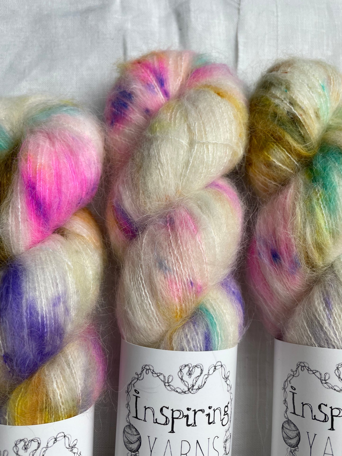 Close up of three skeins of brightly speckled natural coloured hand dyed fluffy Alpaca Silk Lace yarn labelled Inspiring Yarns on a white linen background.