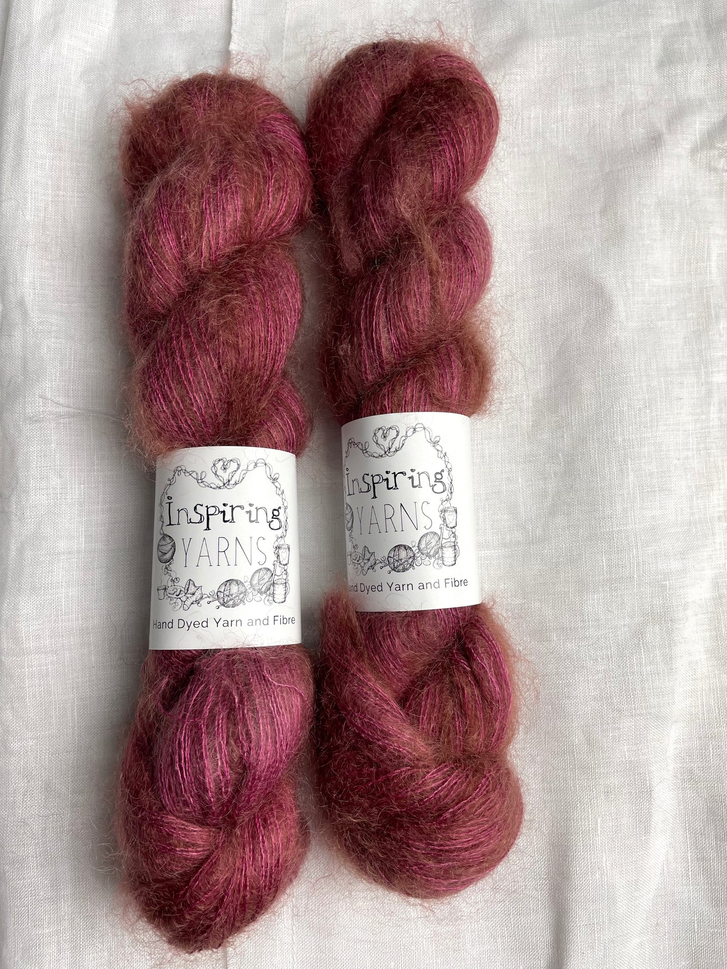 Two skeins of dusky pink mohair silk lace yarn labelled Inspiring Yarns on a white linen background.