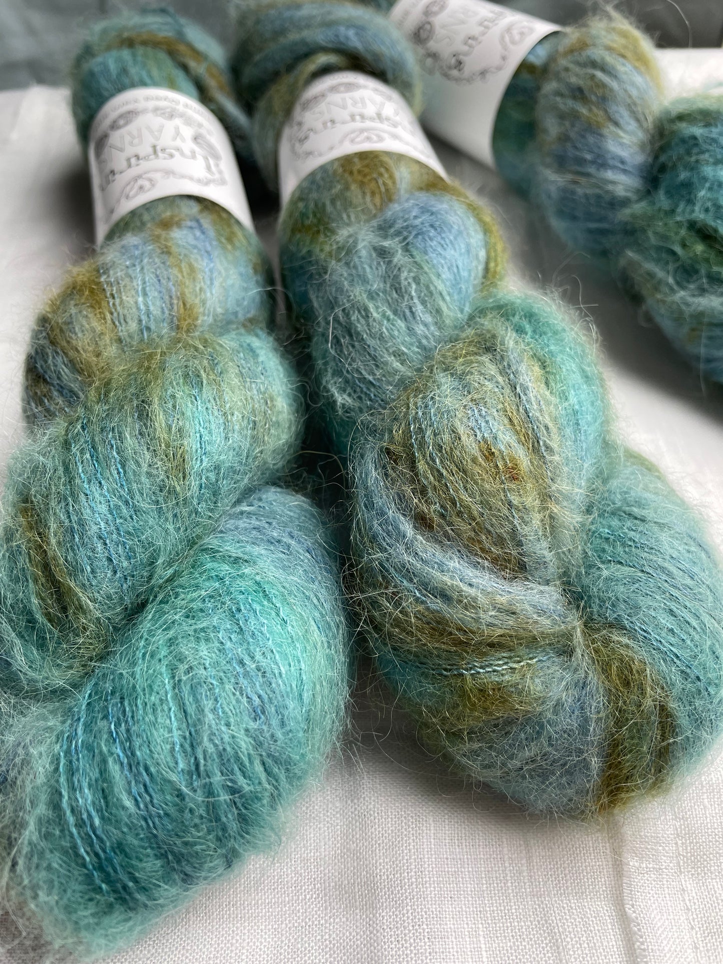 Close up of three skeins of speckled deep teal coloured hand dyed fluffy Alpaca Silk Lace yarn labelled Inspiring Yarns on a white linen background.