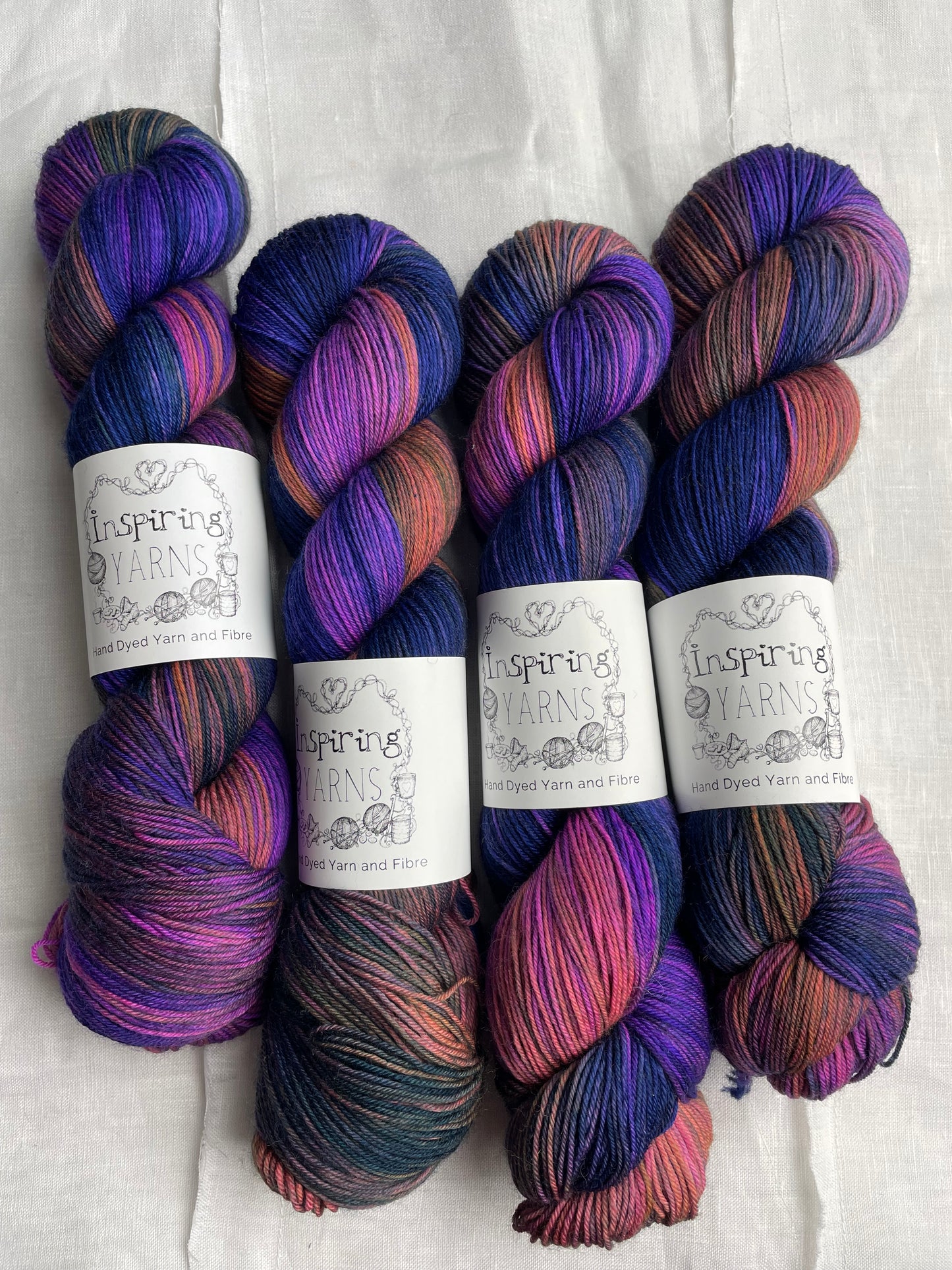 Four variegated blue and multi coloured skeins of Merino Smooth Sock yarn branded Inspiring Yarns on a white linen fabric background.