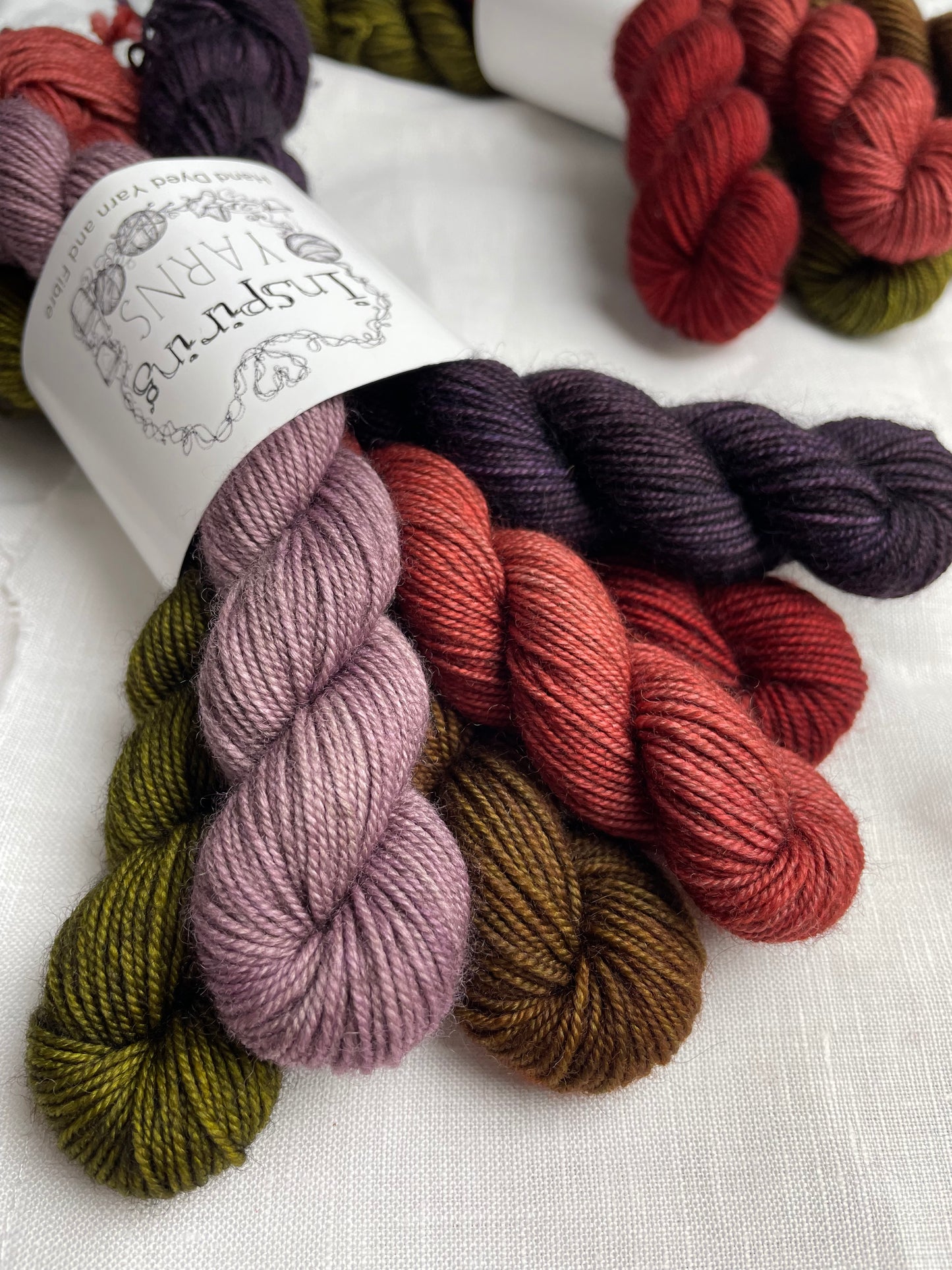 Close up of a set of 6 mini skeins of various colours of Yak Sock yarn labelled Inspiring Yarns on a white linen background.