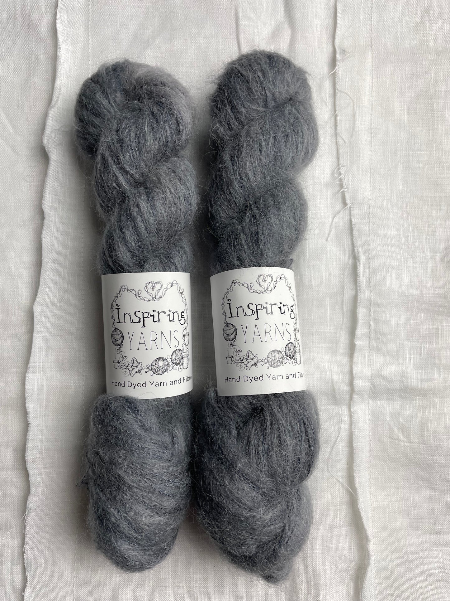 Two skeins of tonal charcoal grey coloured hand dyed fluffy Alpaca Silk Lace yarn labelled Inspiring Yarns on a white linen background.