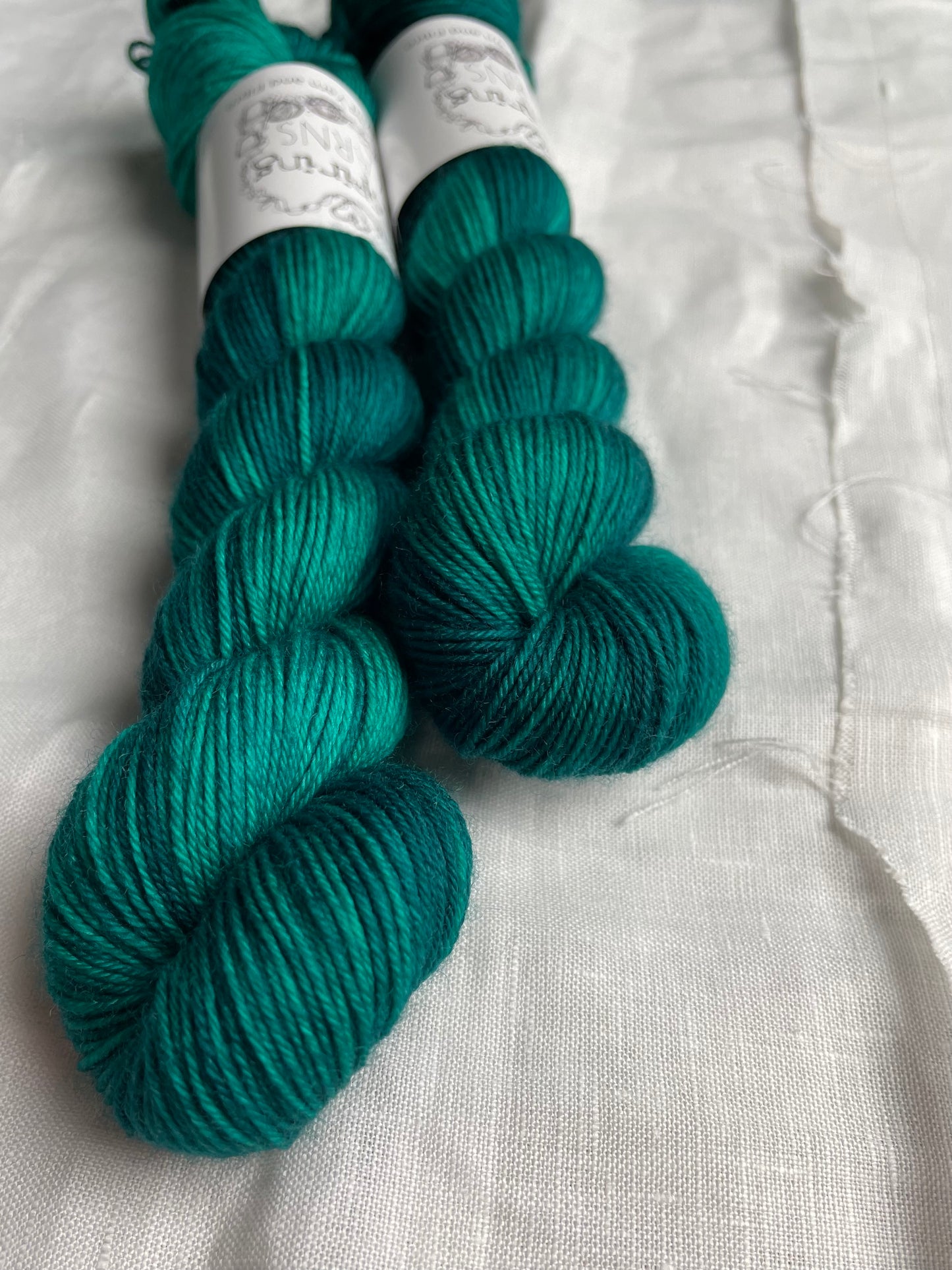 Two skeins of Merino Smooth Sock yarn hand dyed in a tonal jade green colourway and labelled Inspiring Yarns on a white linen background.