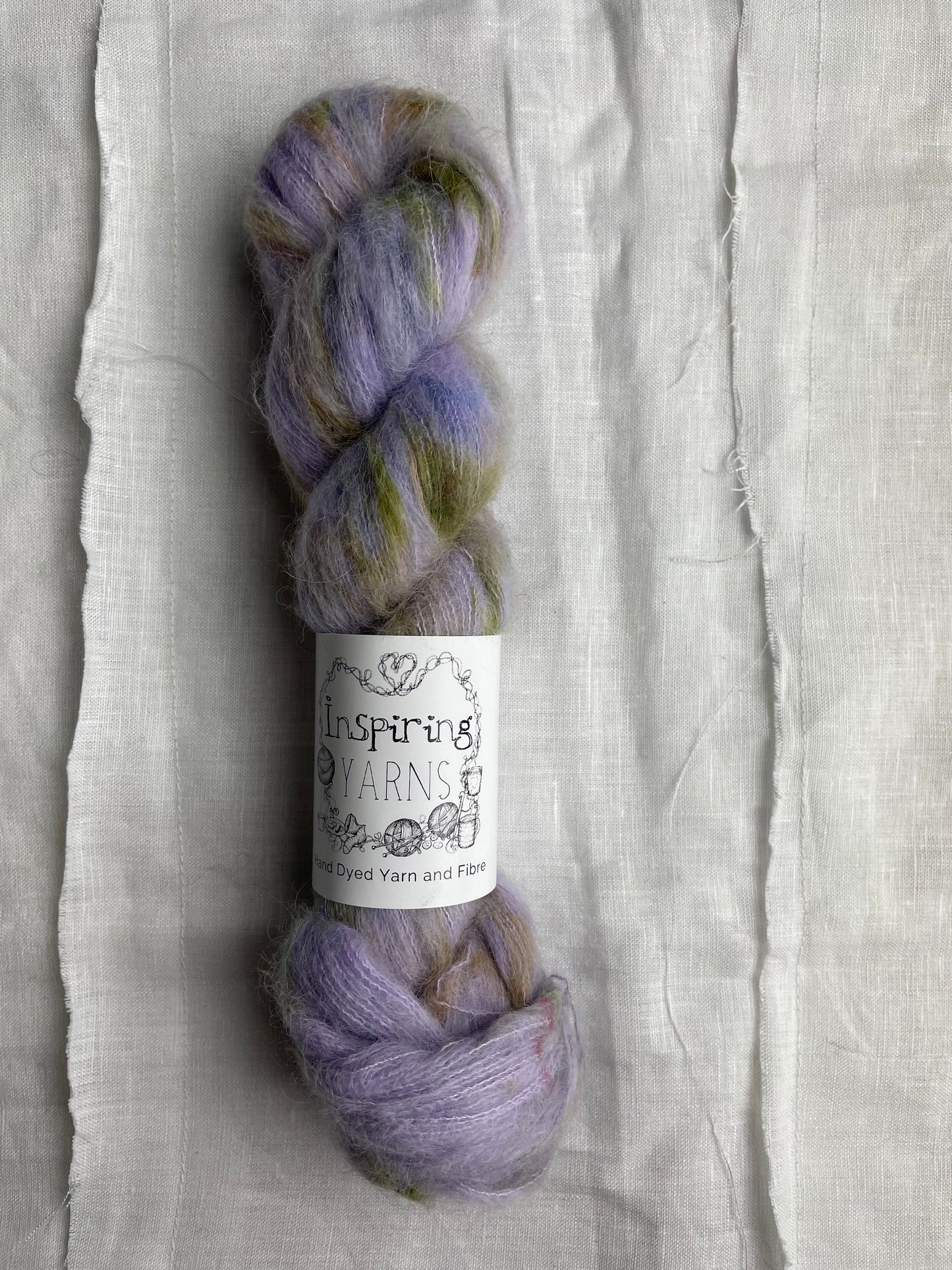 A skein of speckled lilac coloured hand dyed fluffy Alpaca Silk Lace yarn labelled Inspiring Yarns on a white linen background.