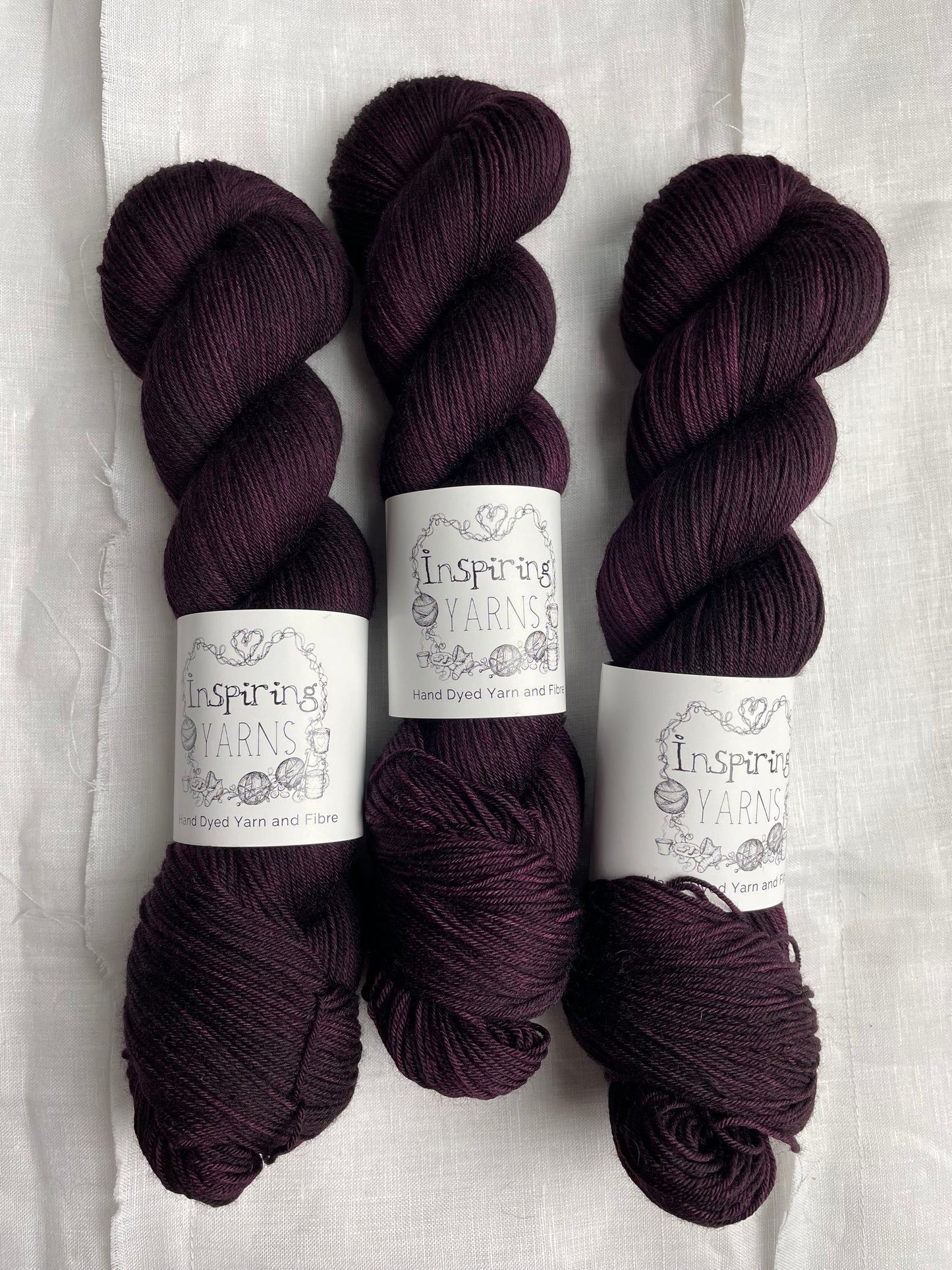 Three skeins of merino smooth sock yarn in a tonal dark burgundy colour labelled Inspiring Yarns on a white linen background.