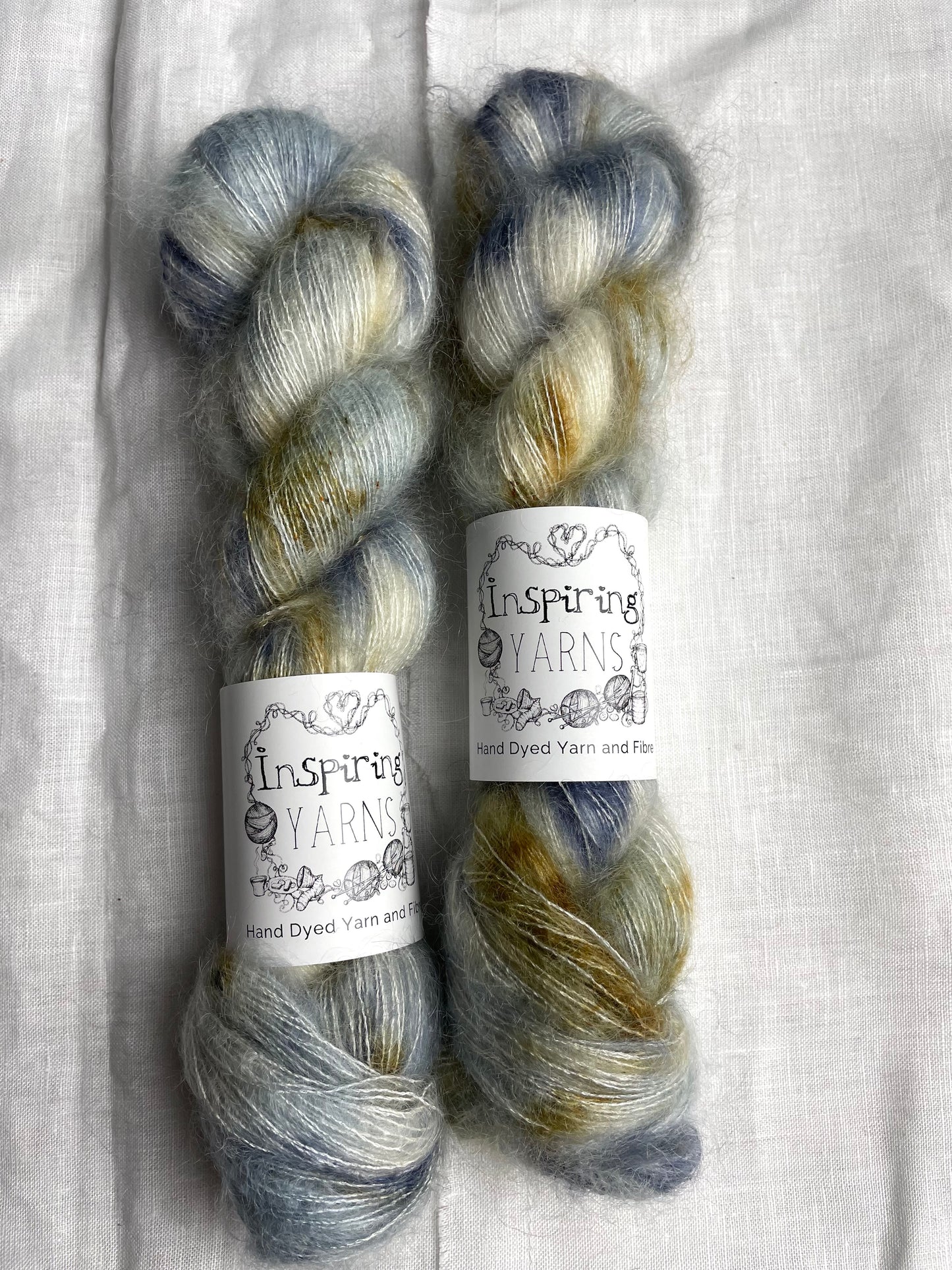 Two skeins of blue and brown speckled mohair silk lace yarn labelled Inspiring Yarns on a white linen background.