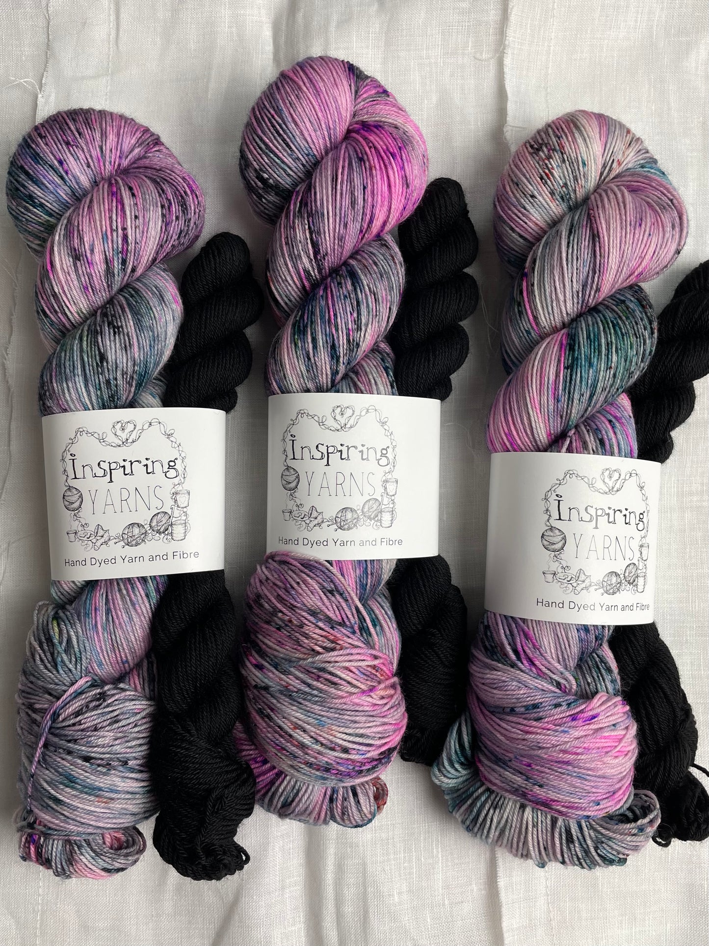 Three sock sets made of a skein of pink black and teal speckled hand dyed yarn with a contrasting black mini skein, labelled Inspiring Yarns and laid on a white linen cloth.