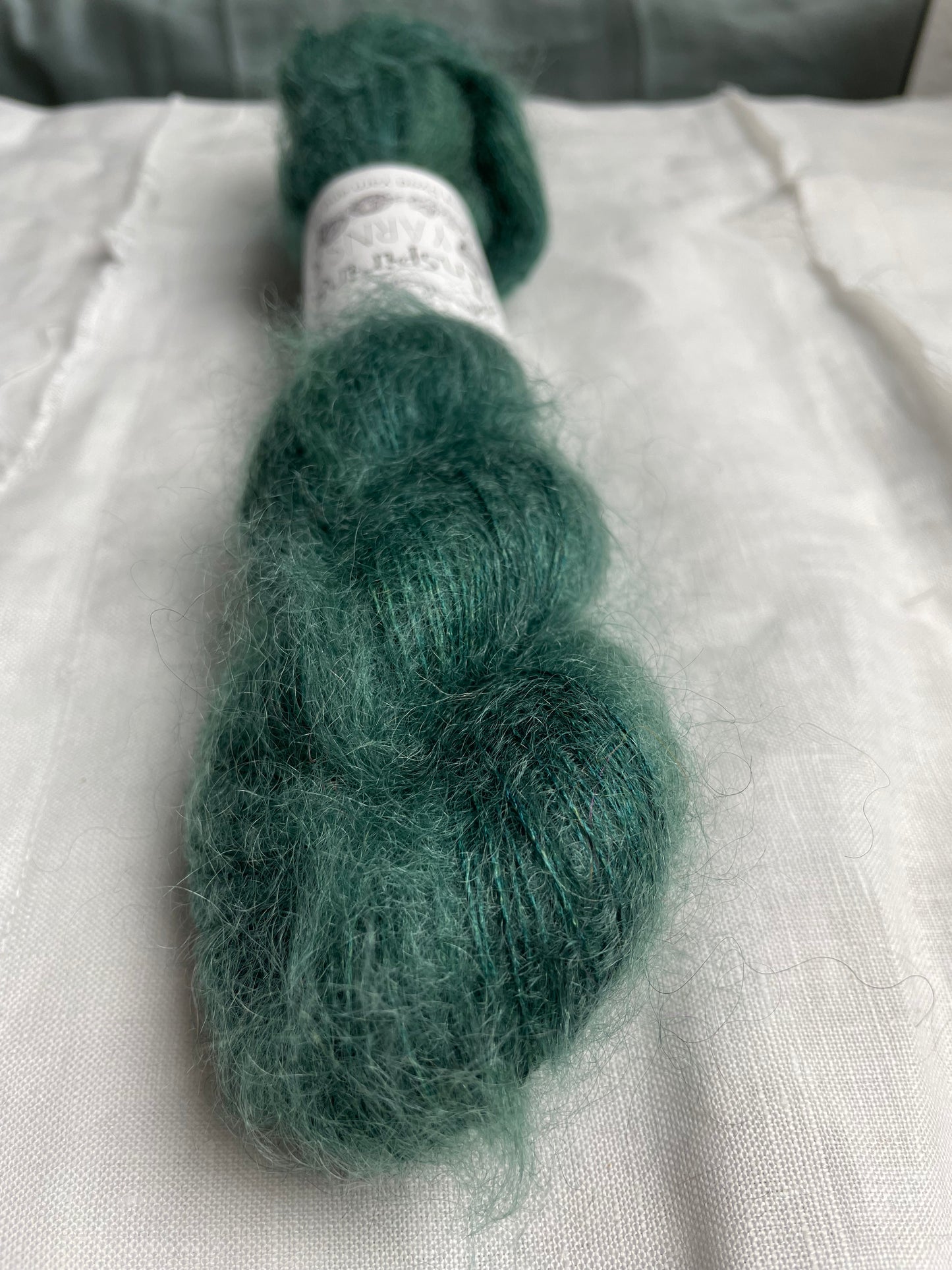 Close up of a skein of tonal teal green kid mohair silk lace yarn labelled Inspiring Yarns on a white linen background.