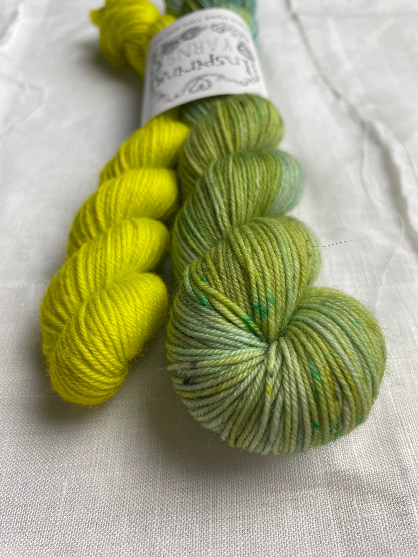 Close up of a set of hand dyed sock yarn comprising one 50g variegated light blue, green and teal and one 20g bright acid yellow on a white linen background. The yarn is branded Inspiring Yarns.