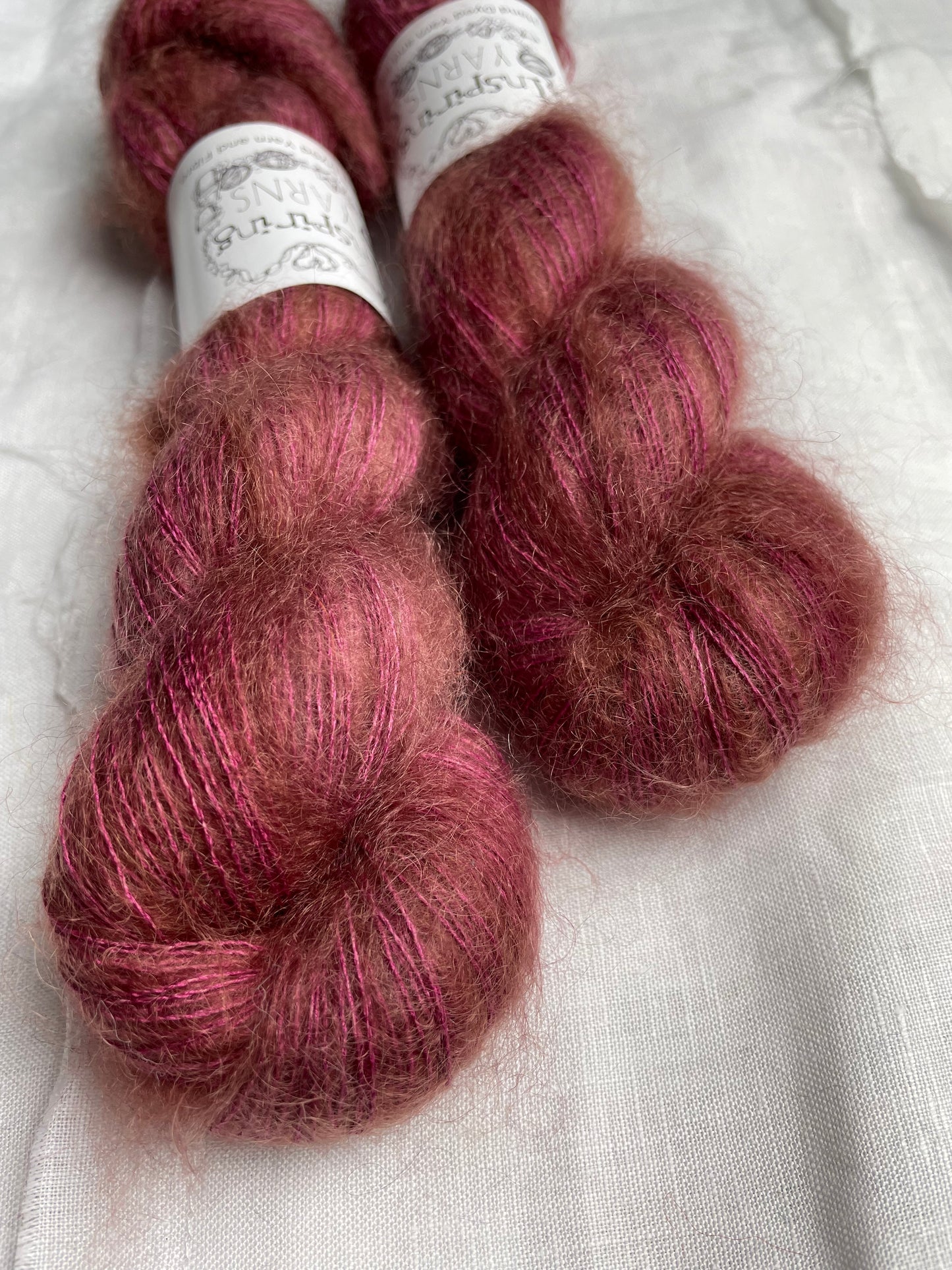 Close up of two skeins of dusky pink mohair silk lace yarn labelled Inspiring Yarns on a white linen background.