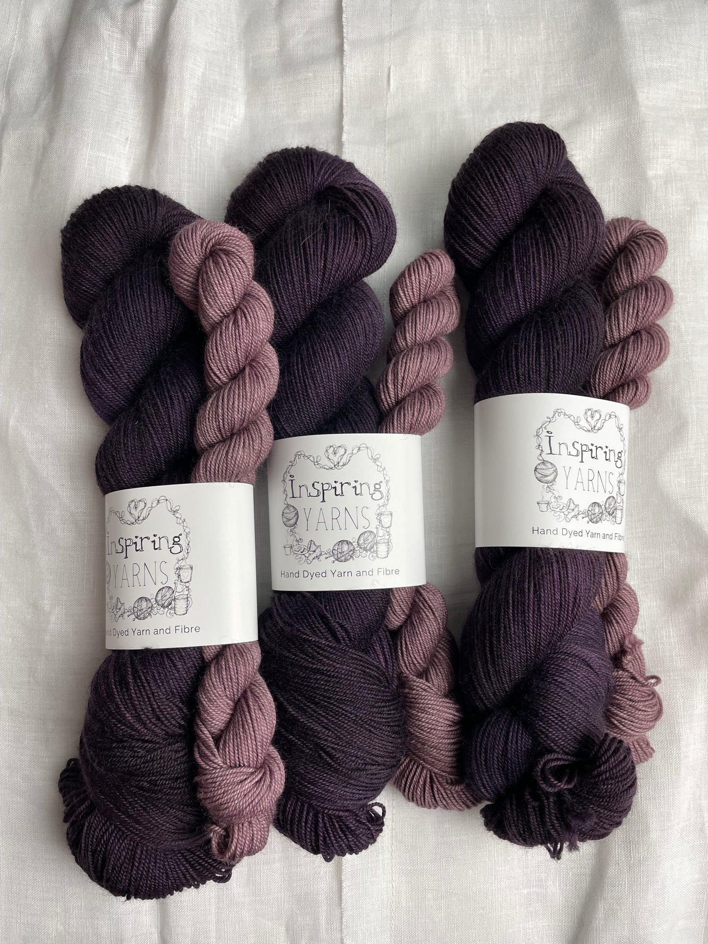 three sock sets made of a skein of dark tonal purple hand dyed yarn with a contrasting dusty lilac mini skein, labelled Inspiring Yarns and laid on a white linen cloth.