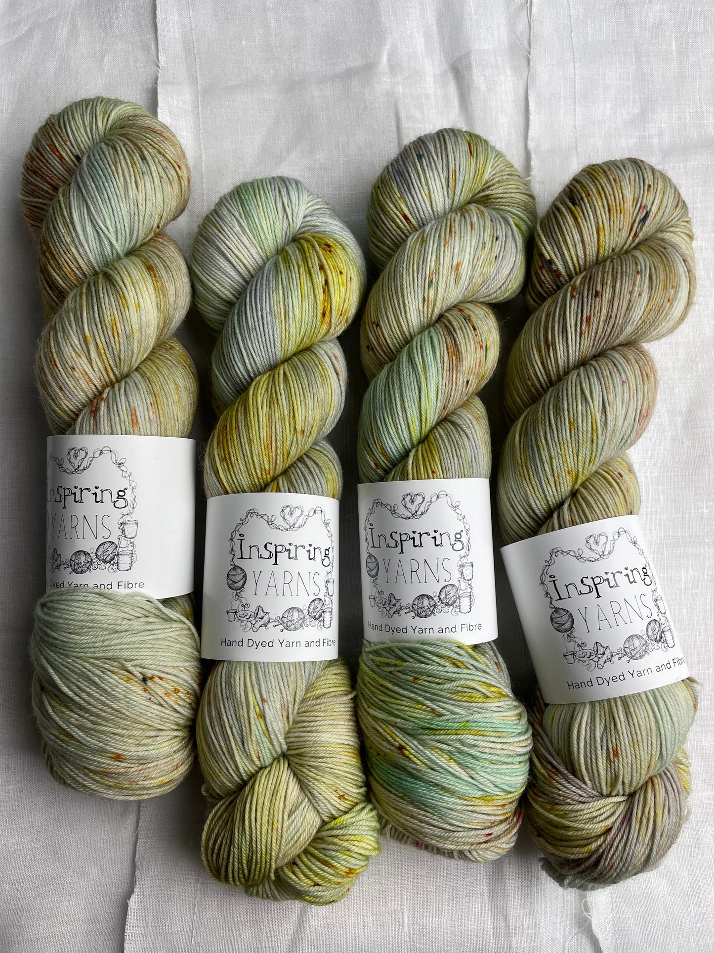 Four lightly speckled, variegated grey and green coloured skeins of Merino Smooth Sock yarn branded Inspiring Yarns on a white linen fabric background.