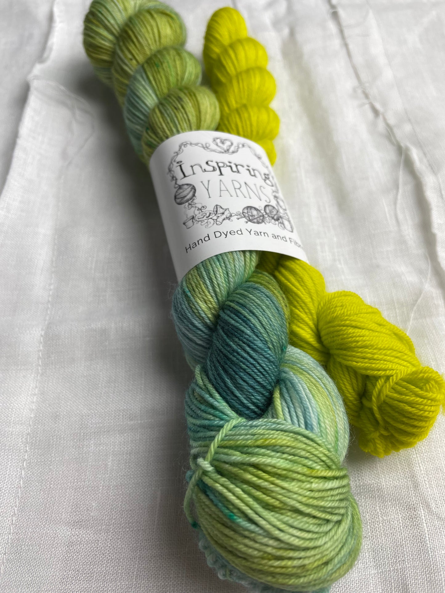 Close up of a set of hand dyed sock yarn comprising one 50g variegated light blue, green and teal and one 20g bright acid yellow on a white linen background. The yarn is branded Inspiring Yarns.