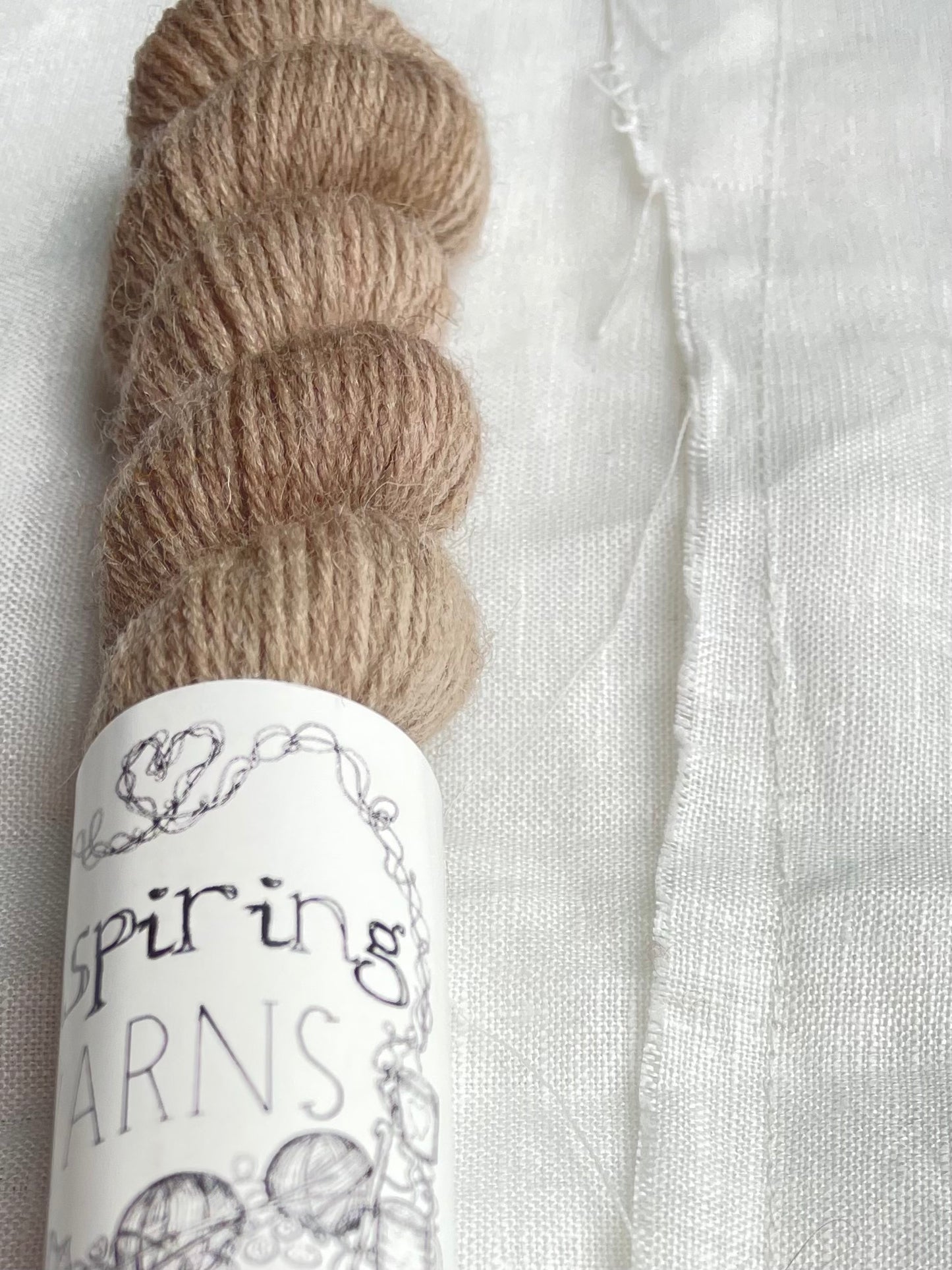 A close up of a mini skein of tonal taupe coloured Highland 4Ply yarn branded Inspiring Yarns on a white linen background.