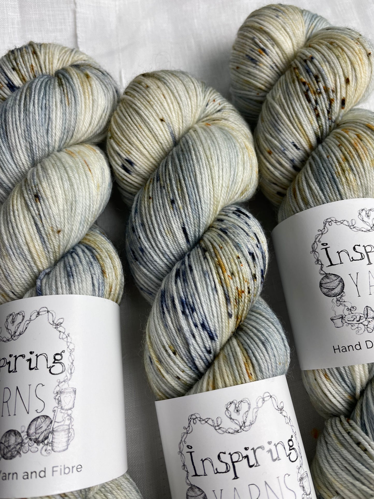 Close up of 3 skeins of blue and brown speckled Merino Smooth Sock yarn labelled Inspiring Yarns on a white linen background.
