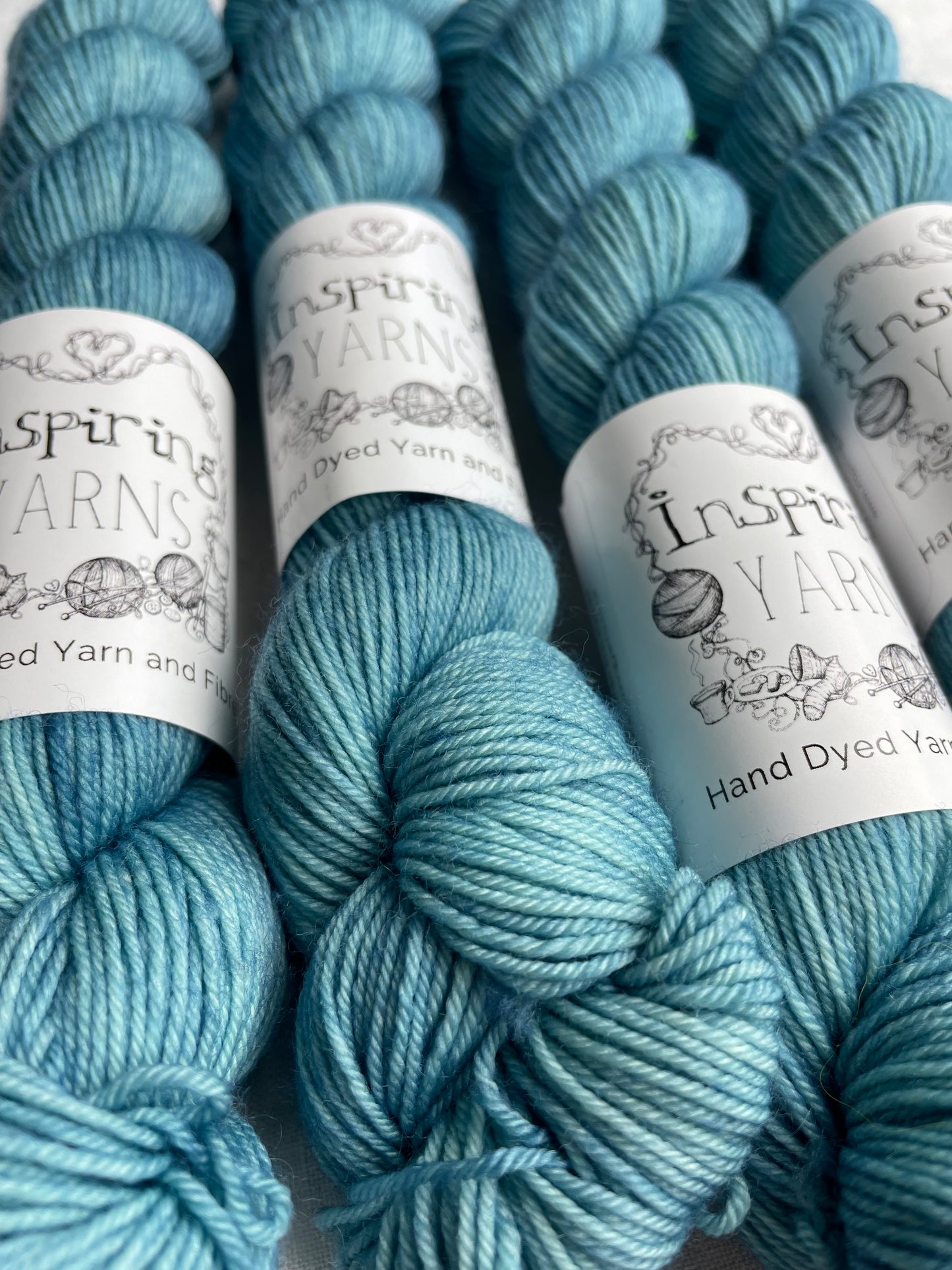 Close up image of four tonal soft teal coloured skeins of hand dyed Merino Smooth Sock yarn branded Inspiring Yarns on white linen fabric.