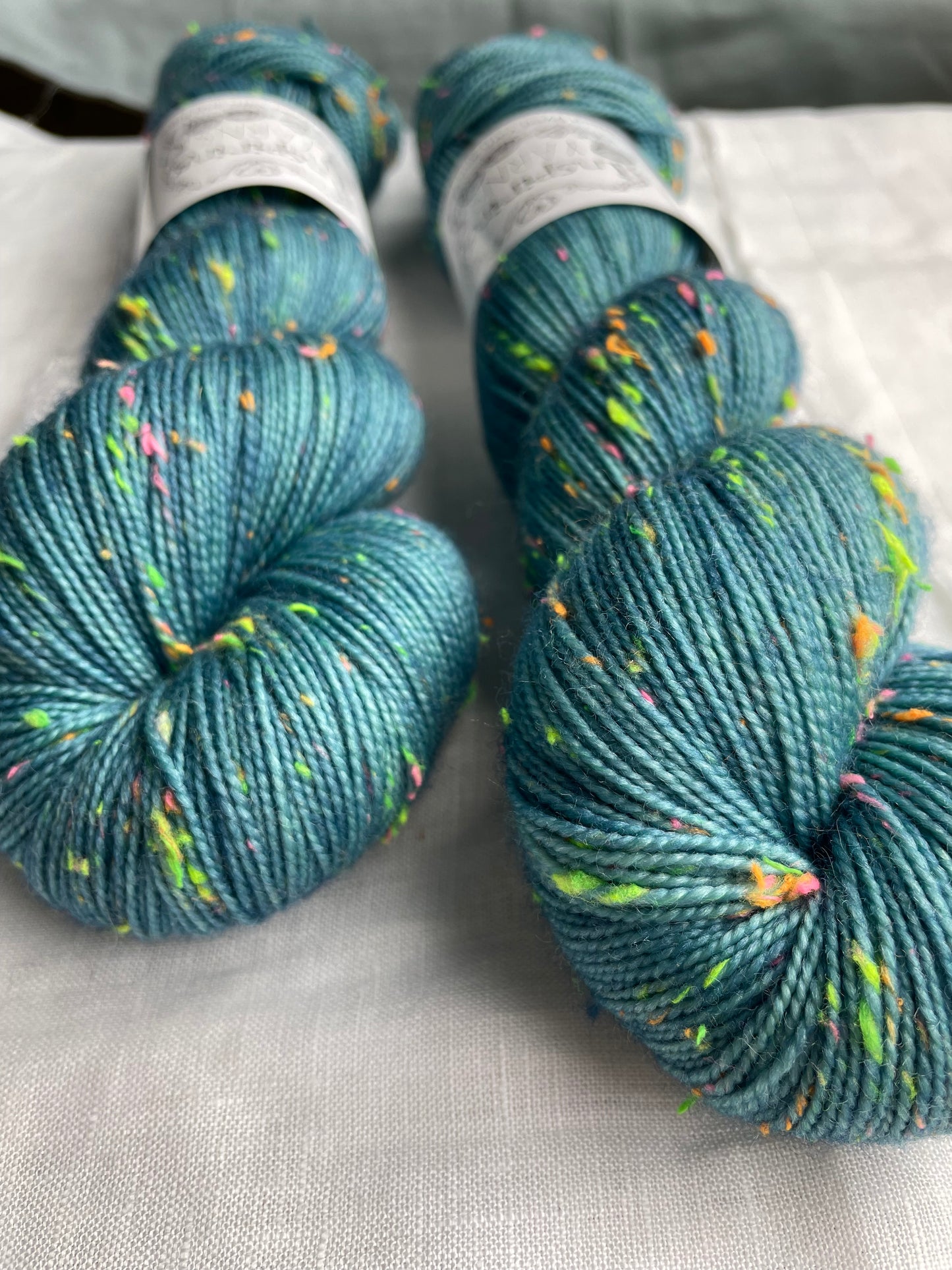 Close up of two skeins of tonal soft teal coloured hand dyed Disco Donegal yarn branded Inspiring Yarns on a white linen background. The yarn has multi coloured neon neps.