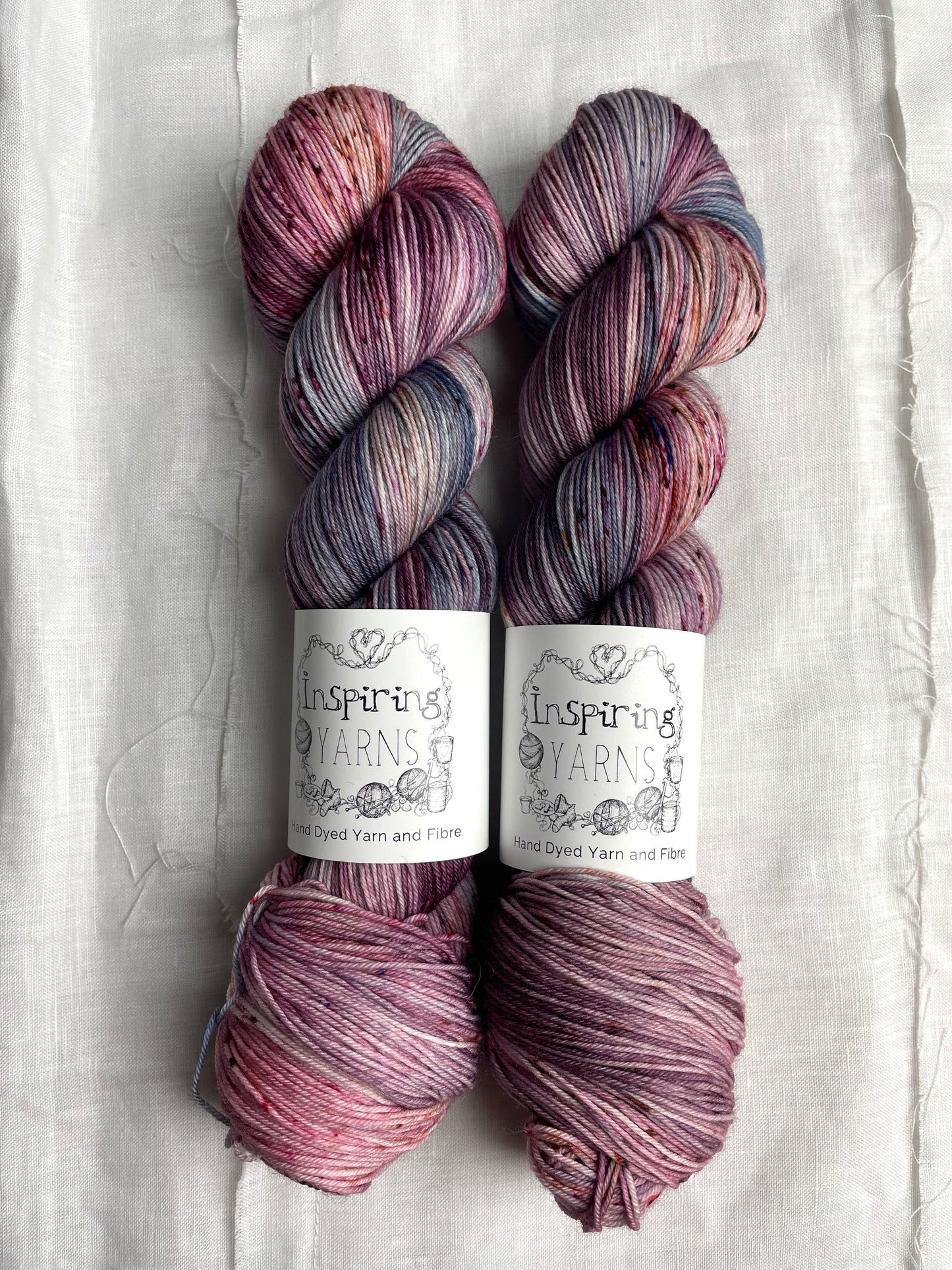 Two skeins of merino smooth sock yarn in shades of pink and blue labelled Inspiring Yarns on a white linen background.