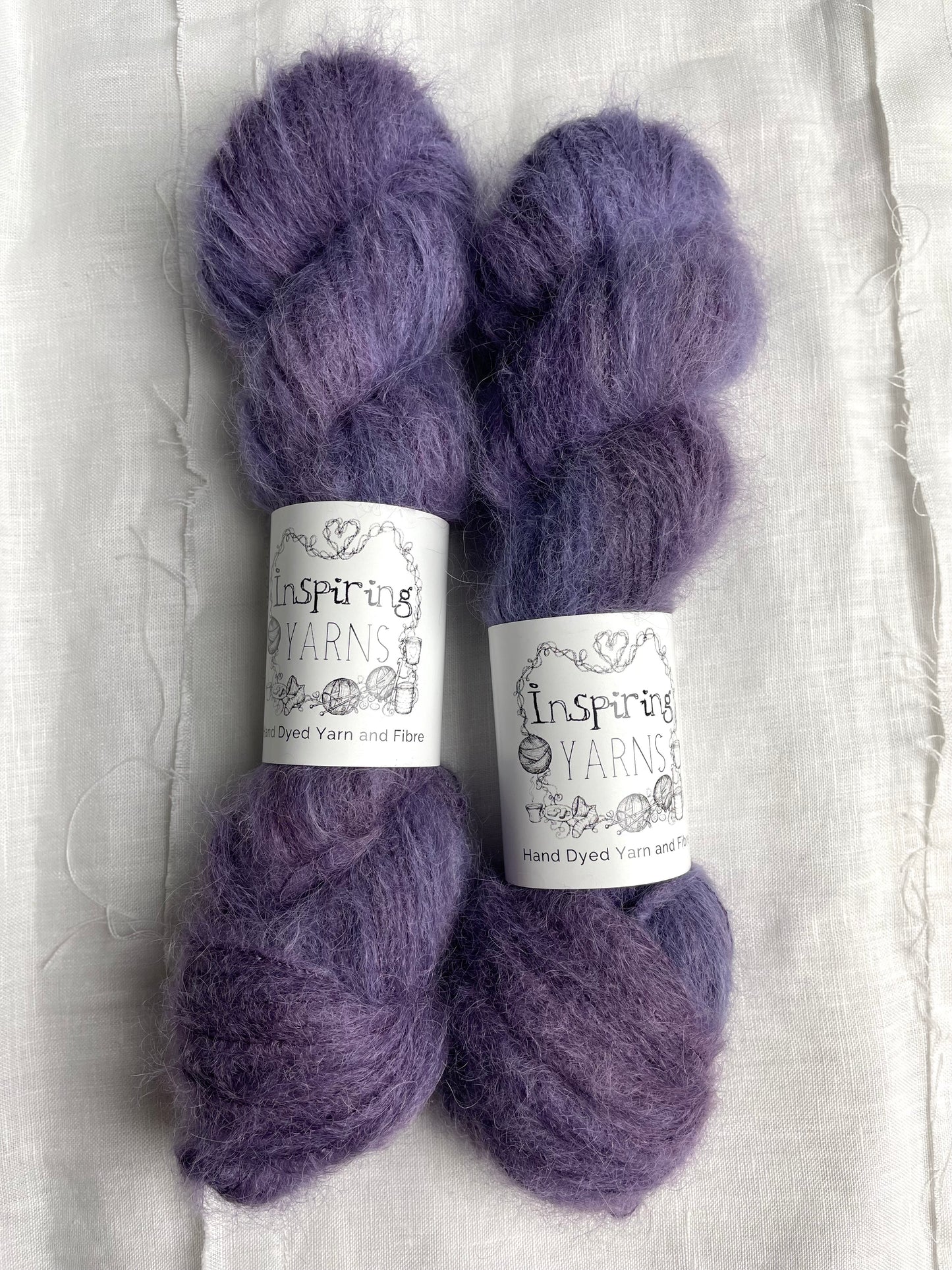 Two skeins of brushed suri silk yarn in a tonal dark purple colour labelled Inspiring Yarns on a white linen background.
