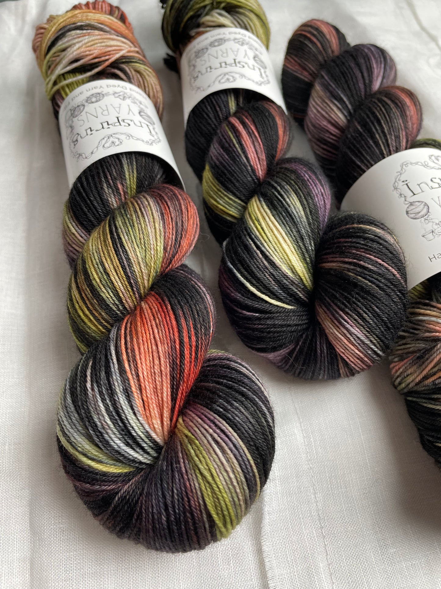 Close up of three skeins of black with lilac green and orange hand dyed yarn, labelled Inspiring Yarns and laid on a white linen cloth.
