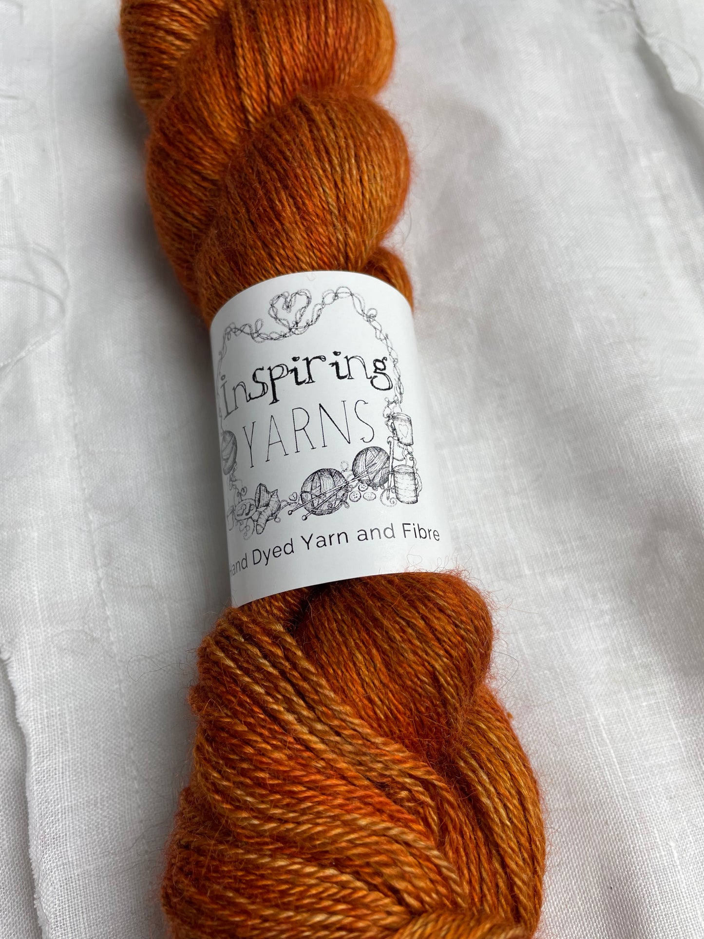 A skein of burnt orange mist 4ply yarn labelled Inspiring Yarns on a white linen background.