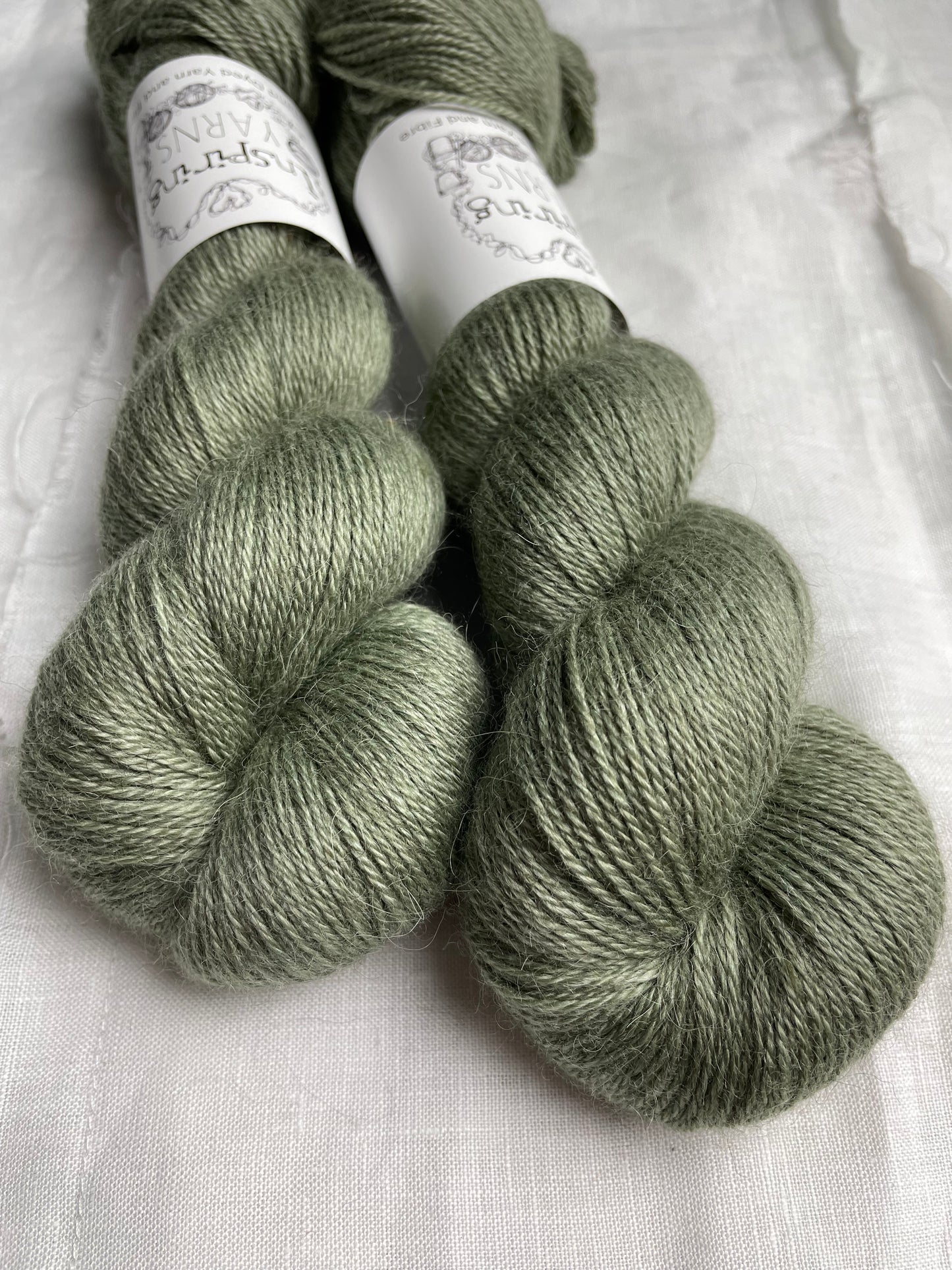 Close up of two skeins of pale green mist 4ply yarn labelled Inspiring Yarns on a white linen background.