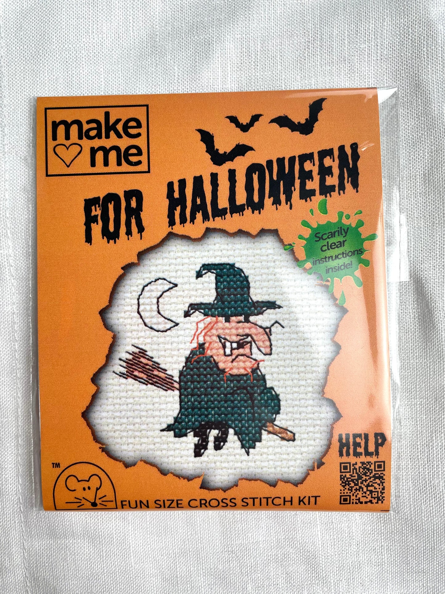 A mini crosstitch kit for a witch on a broomstick with a crescent moon photographed on a white linen background. It is branded Mouseloft Make Me For Halloween in orange packaging.