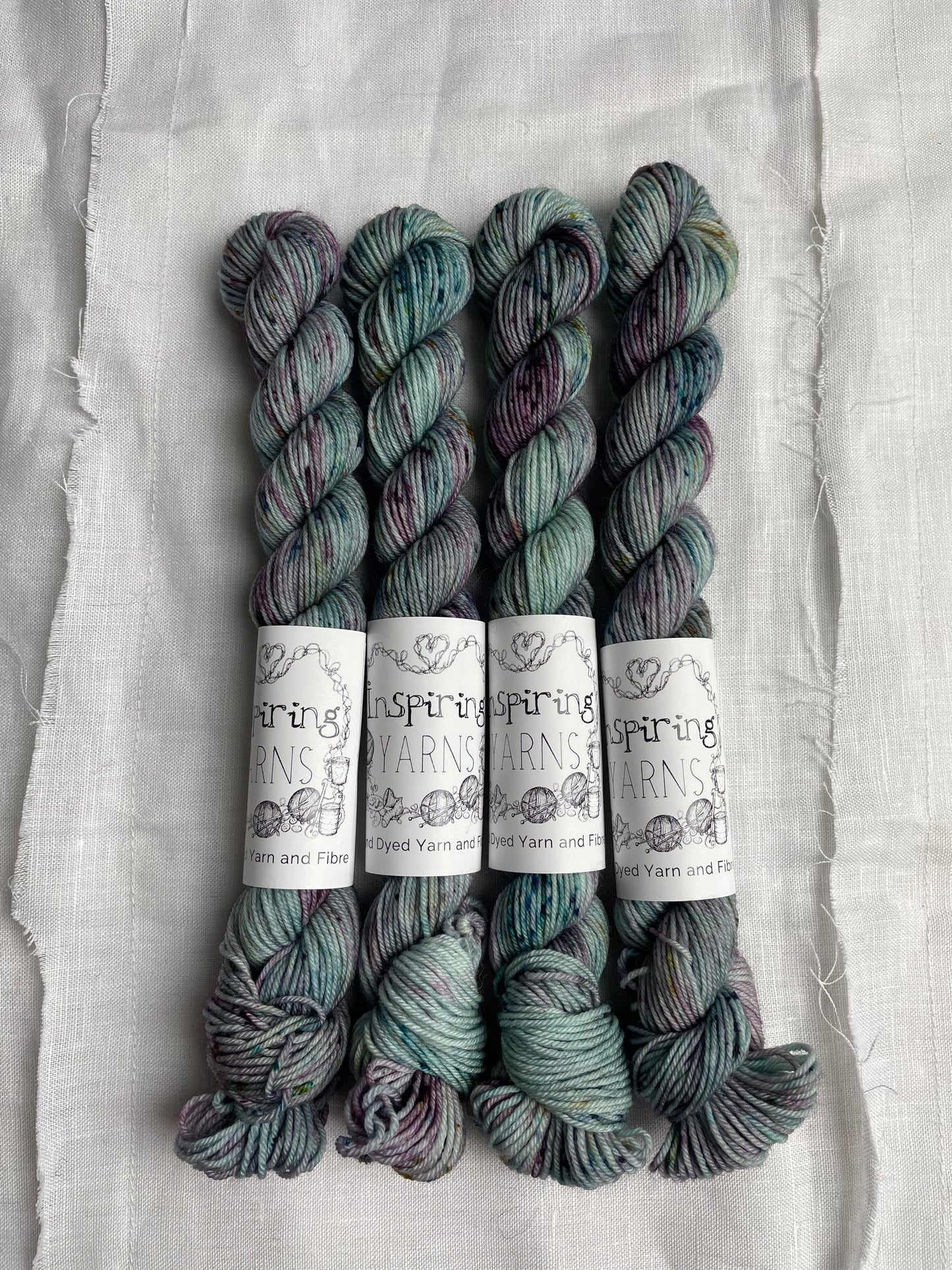 Four skeins of merino smooth sock yarn in a light teal colour with coloured speckles labelled Inspiring Yarns on a white linen background.