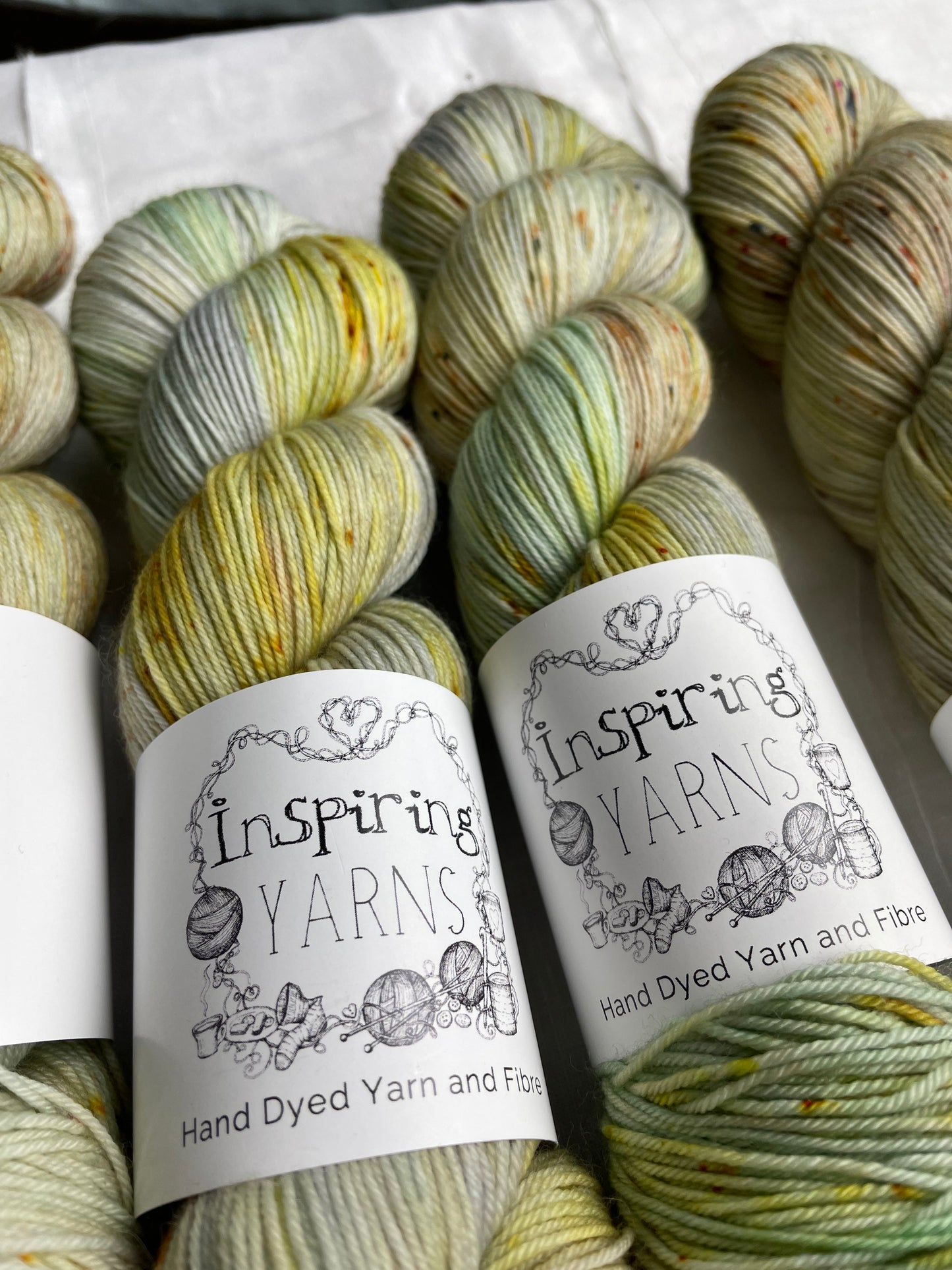 Close up of four lightly speckled, variegated grey and green coloured skeins of Merino Smooth Sock yarn branded Inspiring Yarns on a white linen fabric background.