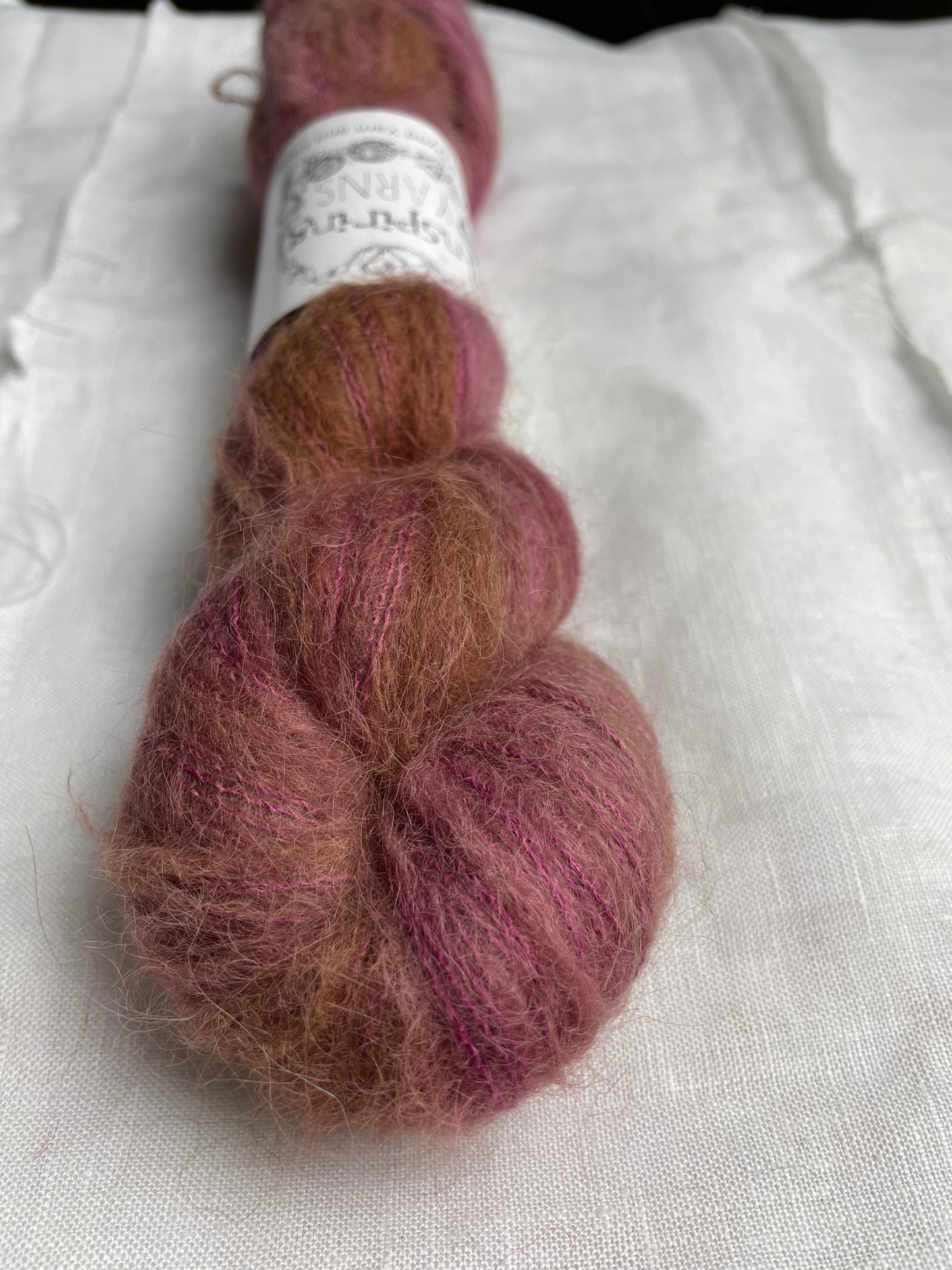 A skein of dusky pink Brushed Baby Suri Silk lace yarn labelled Inspiring Yarns on a white linen background.