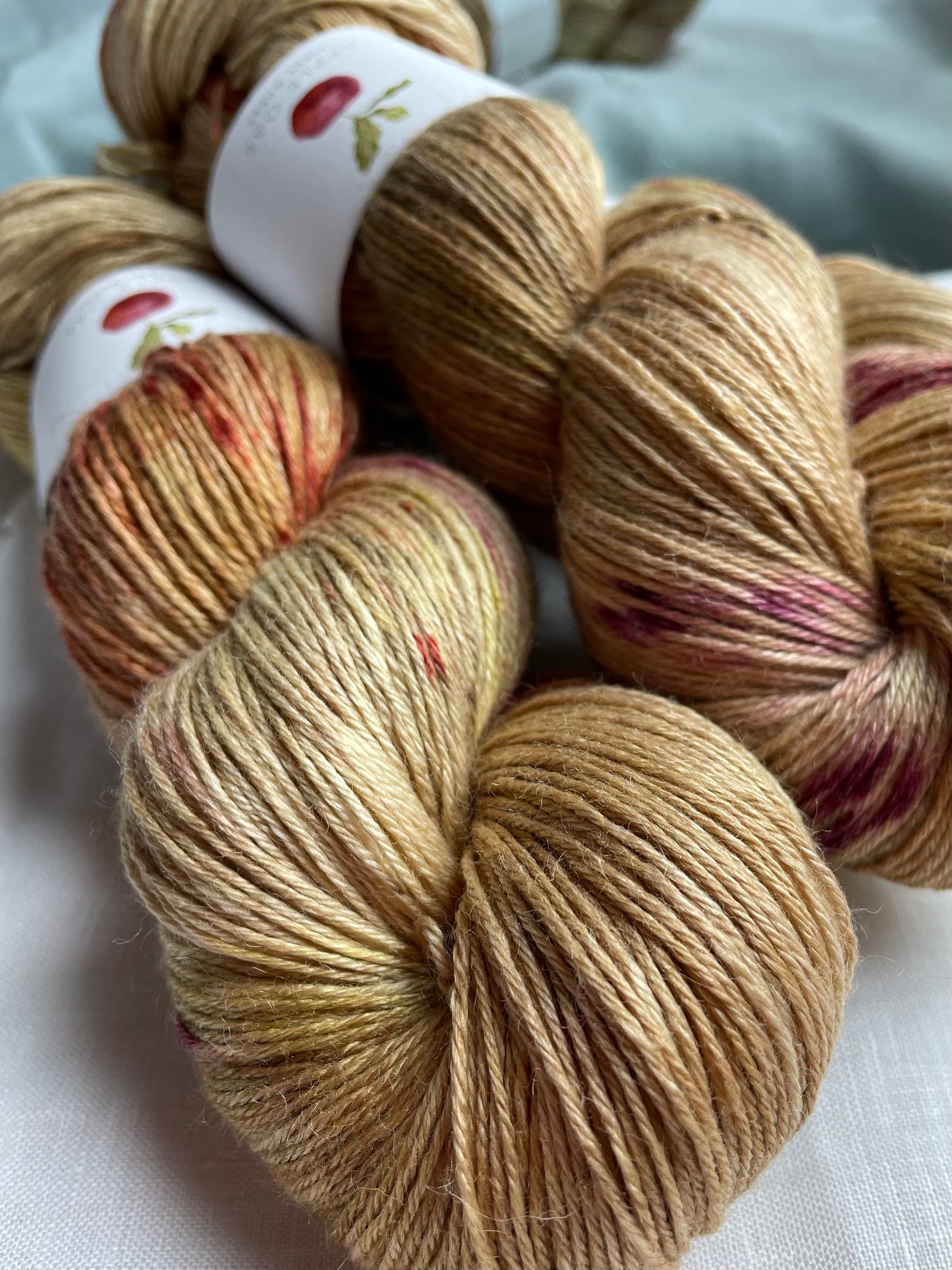 close up of 2 skeins of hand dyed yarn in a golden colour with red and pink splashes, on a white background with a mirror in the background