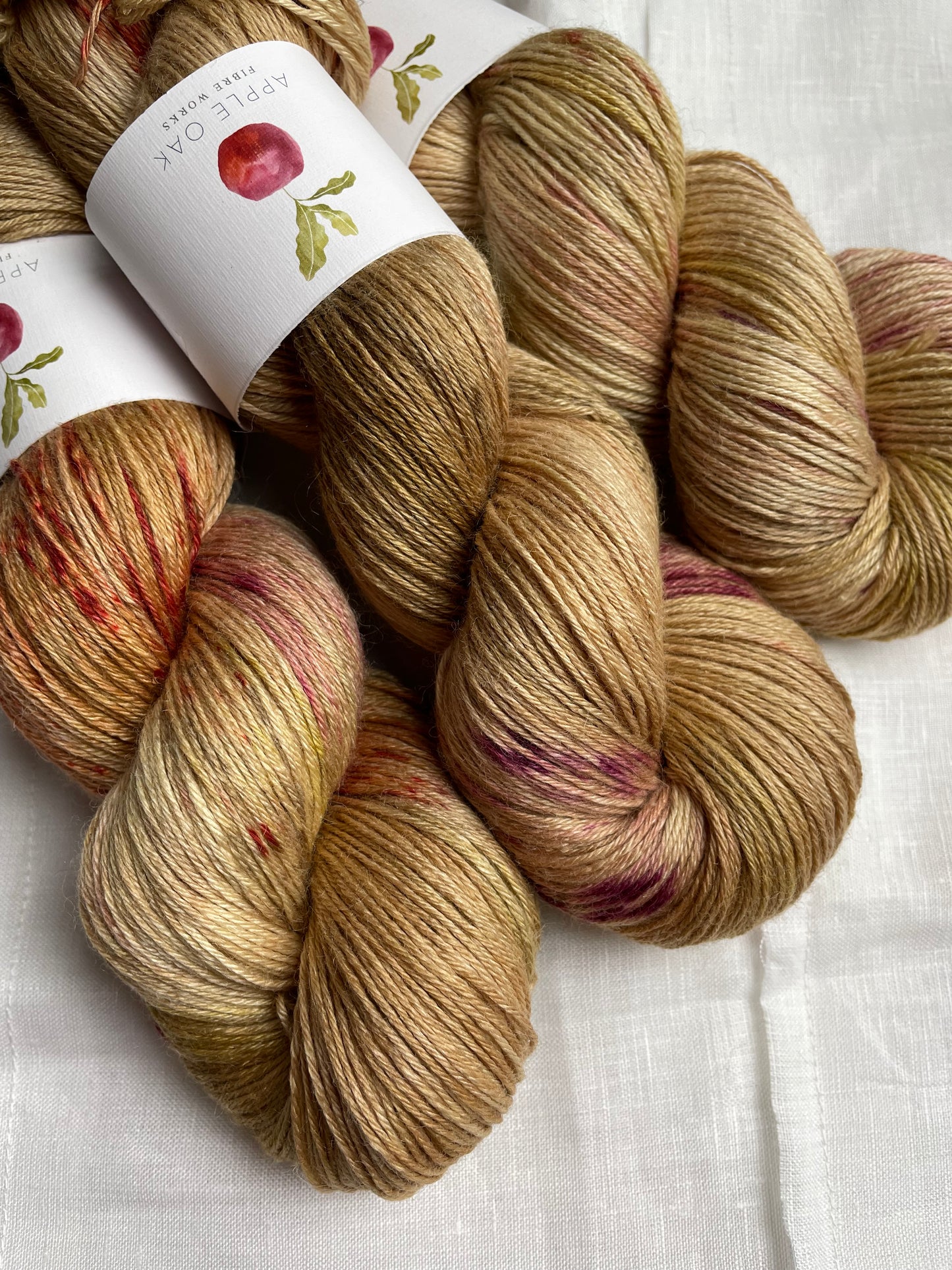 3 skeins of hand dyed yarn in a golden colour with red and pink splashes, the yarn band reads Apple Oak Fibre Works, on a white background