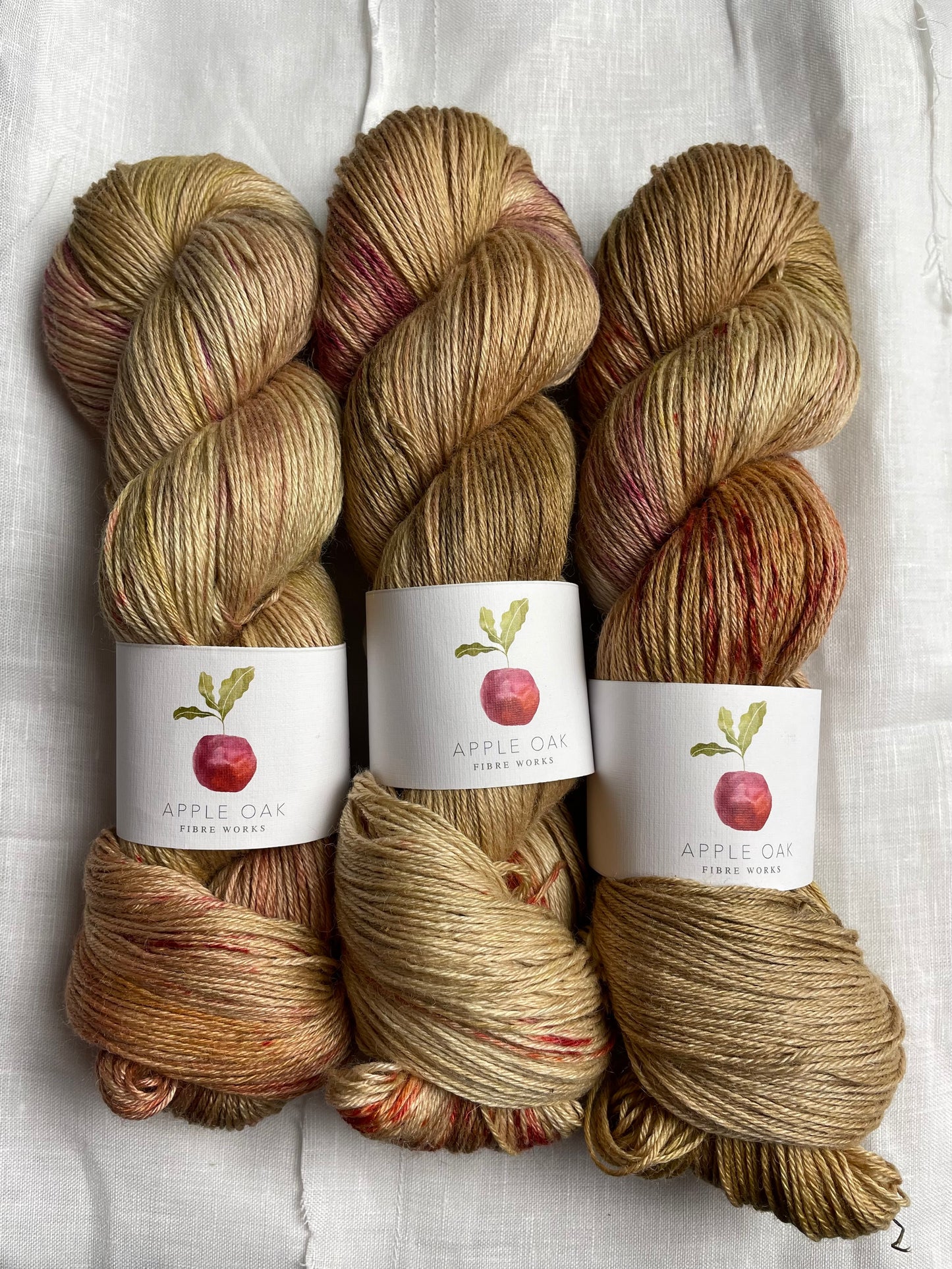 3 skeins of hand dyed yarn in a golden colour with red and pink splashes, the yarn band reads Apple Oak Fibre Works, on a white background