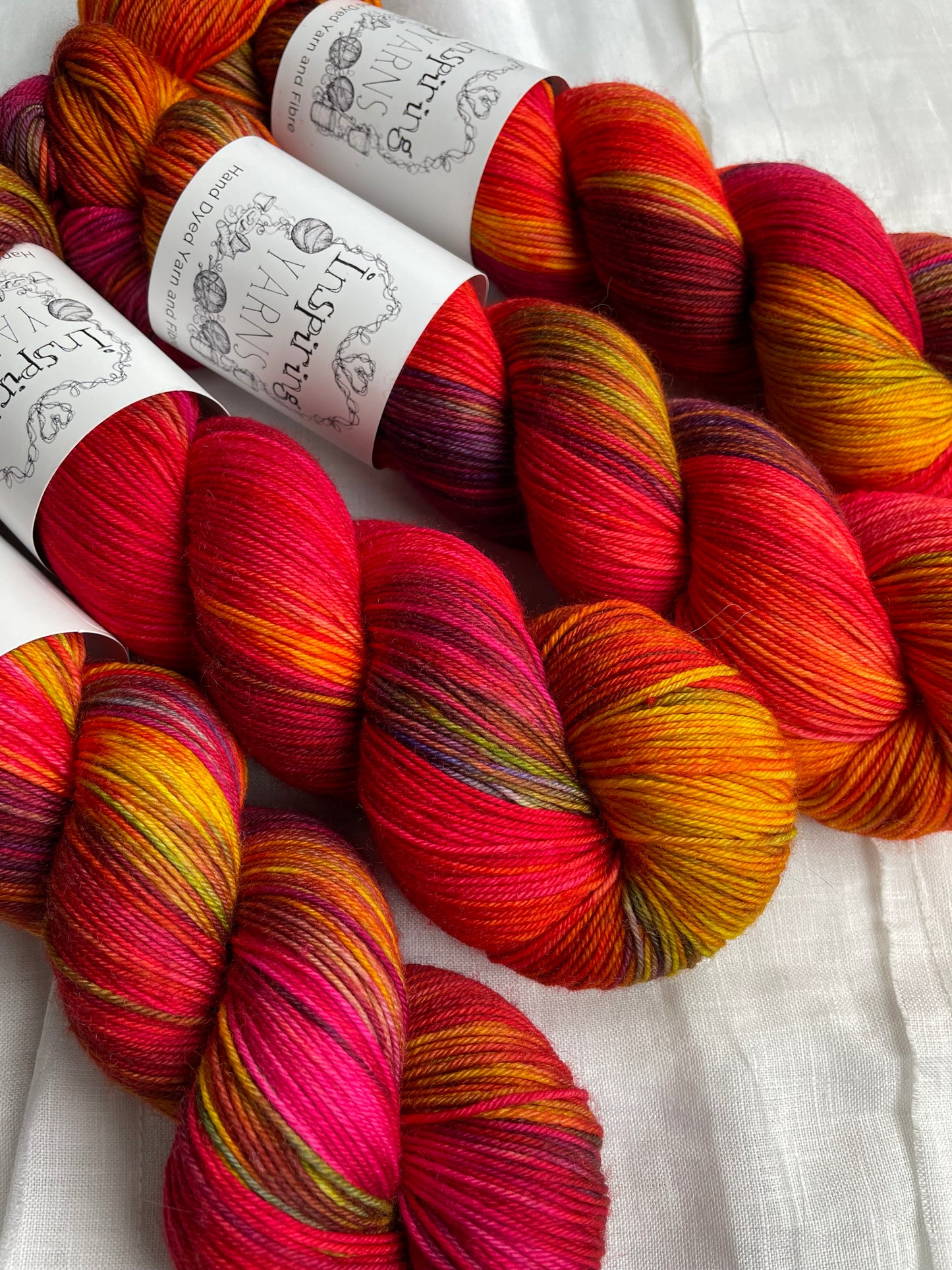 Four variegated red and multi coloured skeins of Merino Smooth Sock yarn branded Inspiring Yarns on a white linen fabric background.
