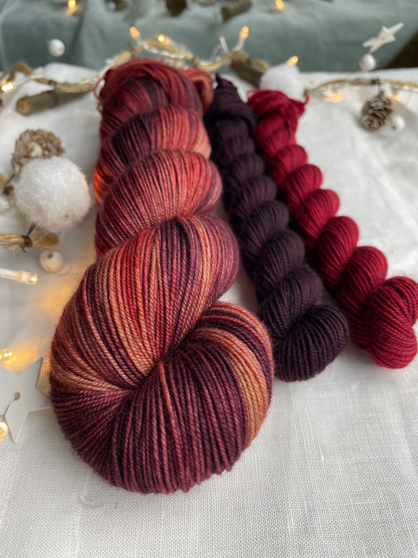 Close up of a hand dyed yarn sock set featuring large skein in burgundy, red and orange tones with a dark red and a burgundy mini skein labelled Inspiring Yarns. The background is white linen and some Christmas lights with pinecones, twigs and stars.