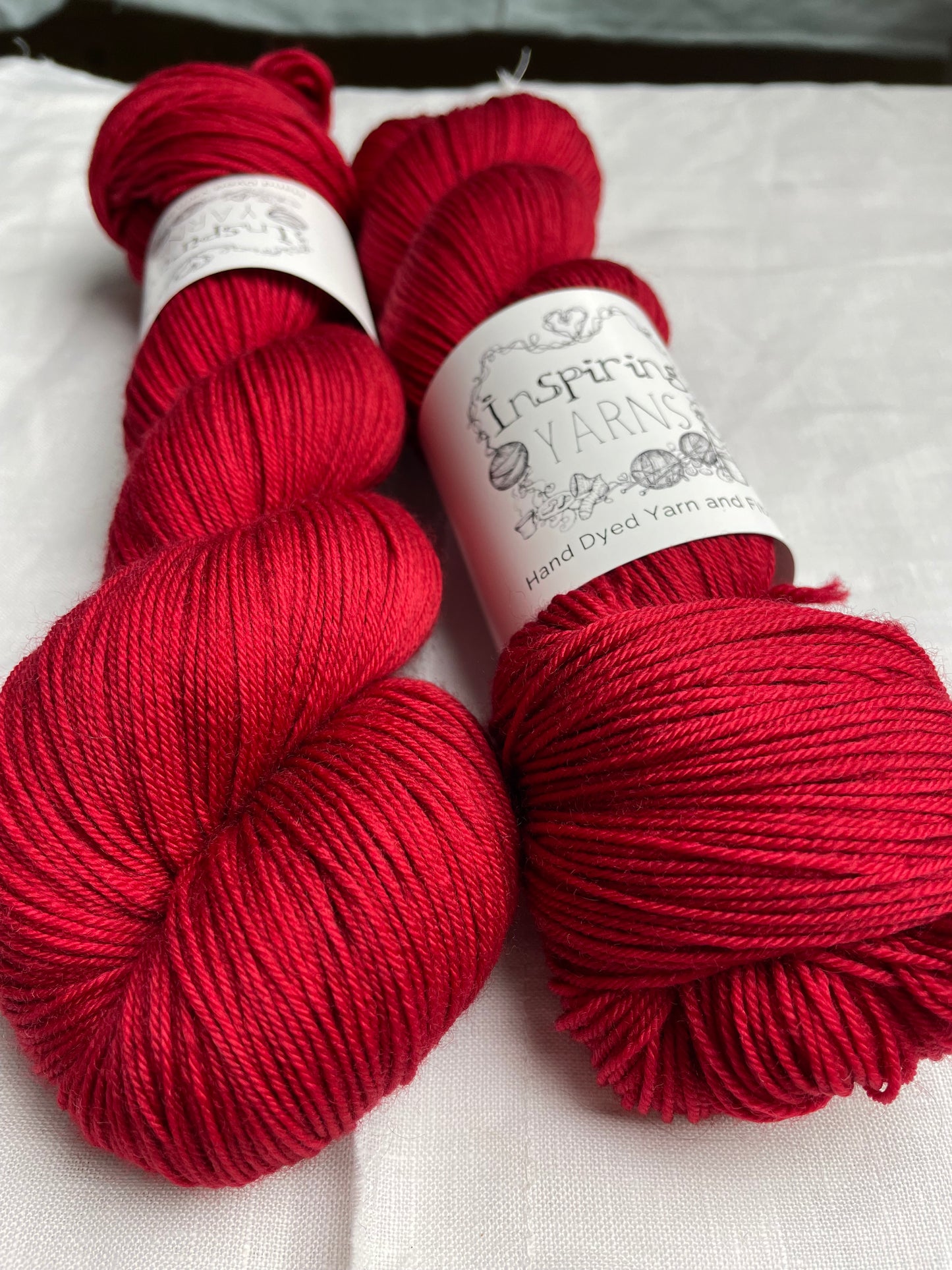 Two skeins of tonal blood red Merino Smooth Sock yarn labelled Inspiring Yarns on a white linen background