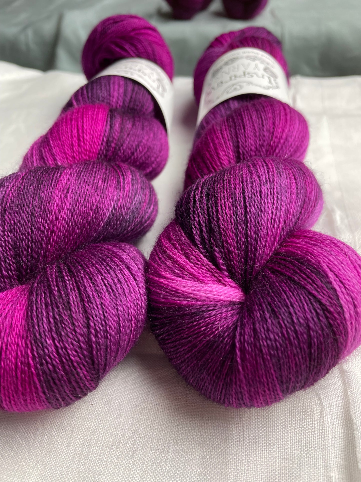 Close up of two skeins of variegated pink plum coloured hand dyed Merino Lace yarn branded Inspiring Yarns on a white linen cloth.