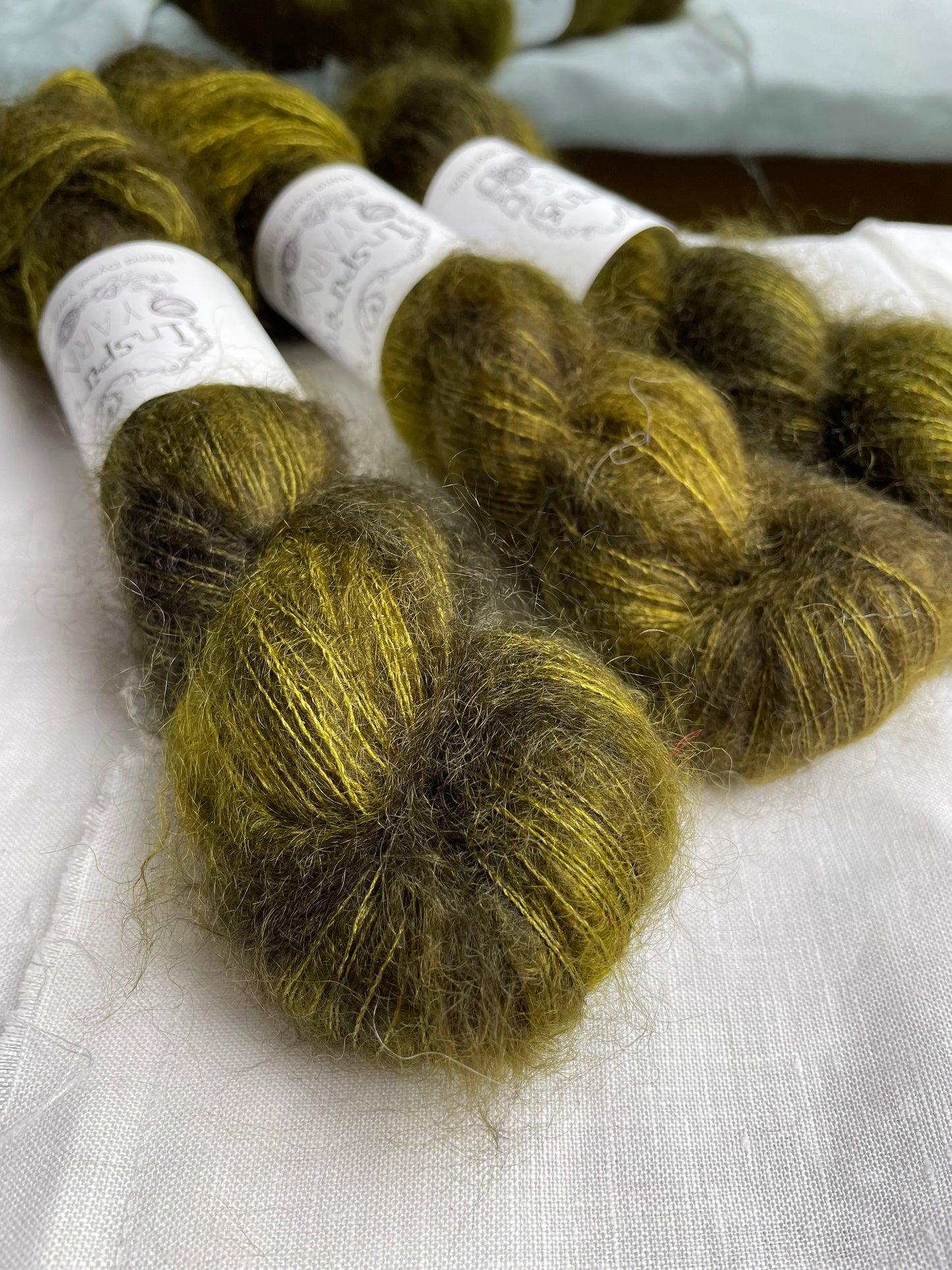 Close up of three skeins of dark olive green mohair silk lace yarn labelled Inspiring Yarns on a white linen background.