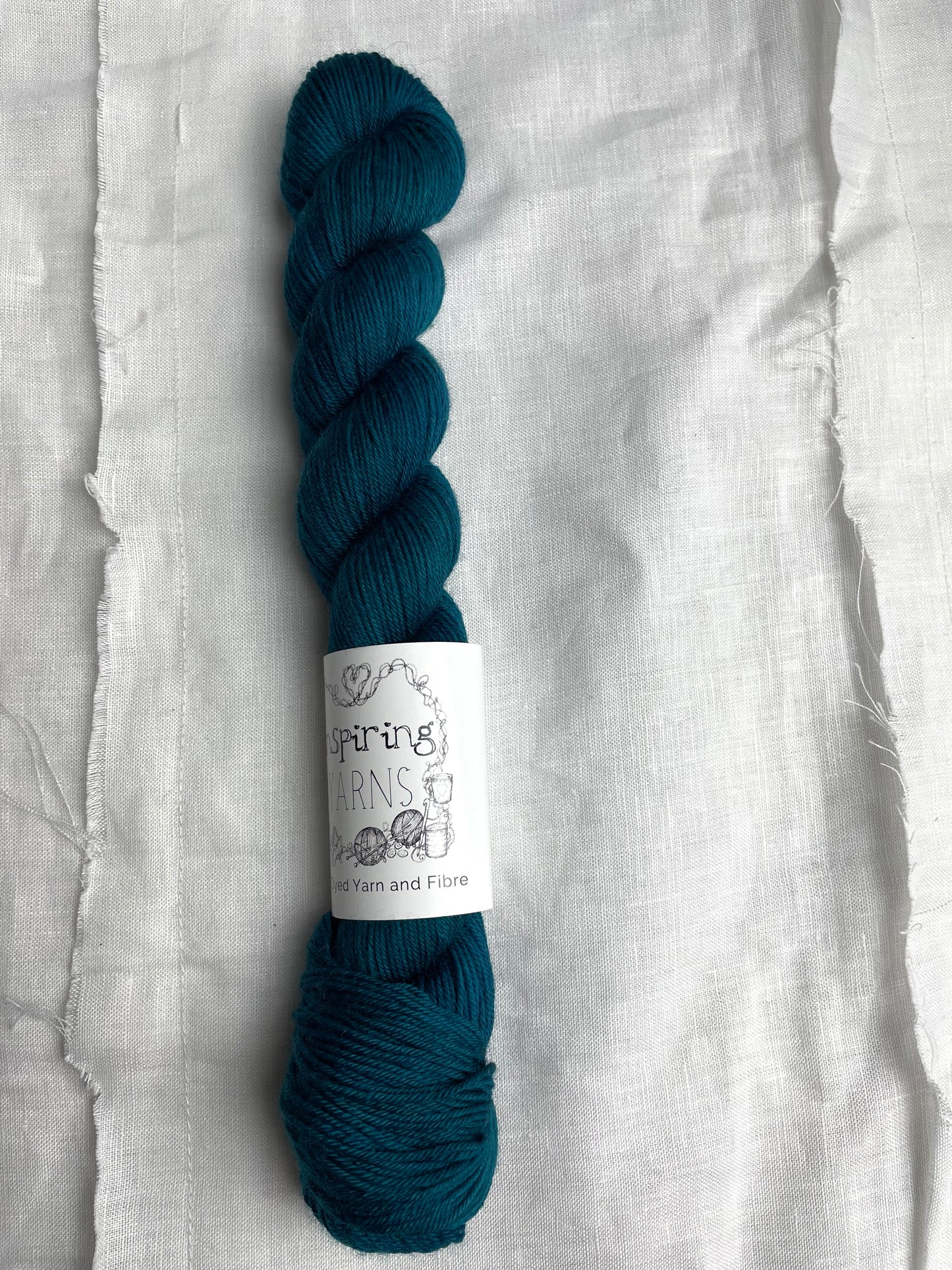 One intense teal blue coloured skeins of Merino Smooth Sock yarn branded Inspiring Yarns on a white linen fabric background.