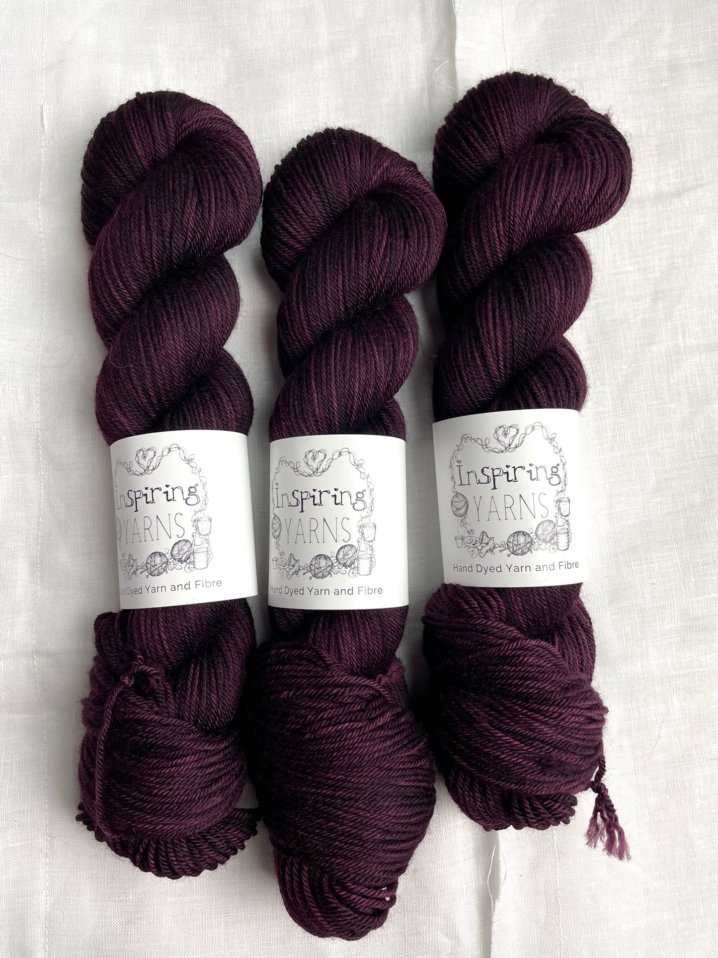 Three skeins of merino smooth DK yarn in a tonal dark burgundy colour labelled Inspiring Yarns on a white linen background.