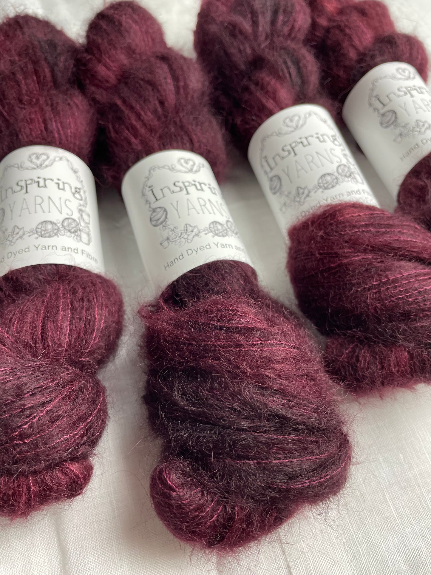 Four skeins of fluffy brushed suri silk lace yarn in a tonal dark burgundy colour labelled Inspiring Yarns on a white linen background.
