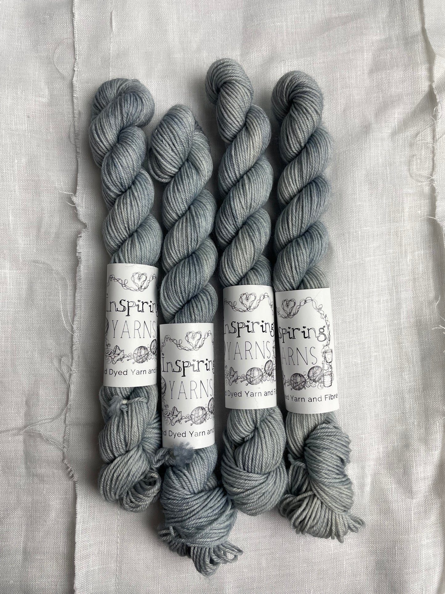 Four mini skeins of Ghost, a pale grey blue coloured Merino Smooth Sock, labelled Inspiring Yarns on a white linen background.