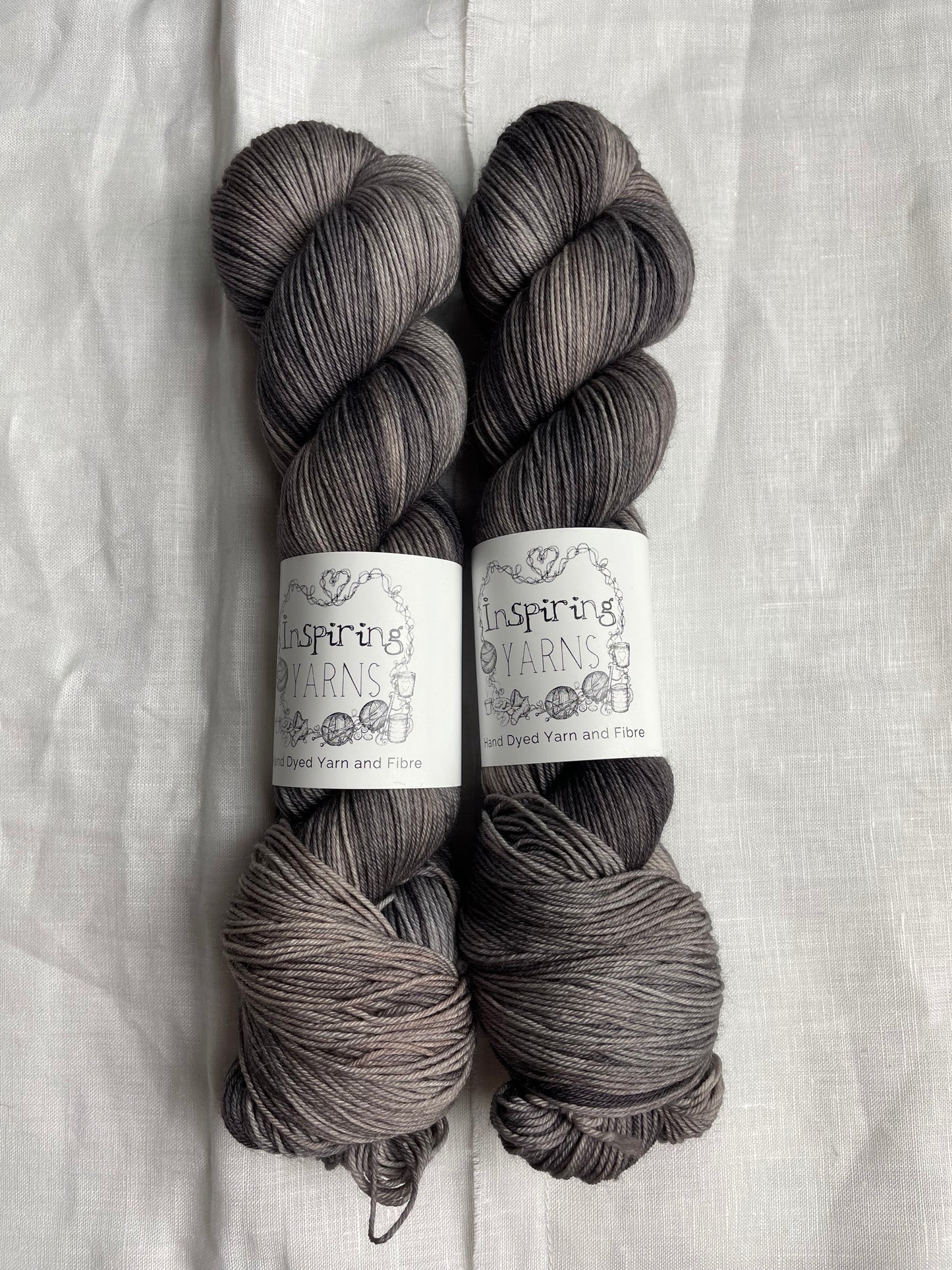 Two skeins of Merino Smooth Sock yarn hand dyed in a tonal grey colourway and labelled Inspiring Yarns on a white linen background.