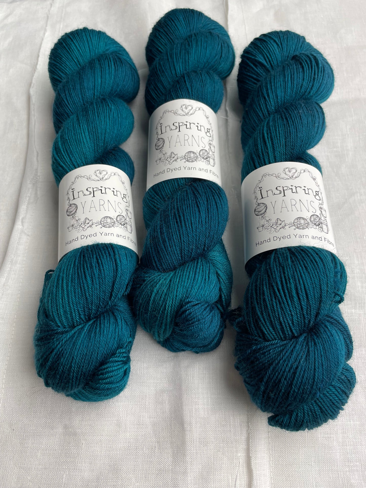 Three intense teal blue coloured skeins of Merino Smooth Sock yarn branded Inspiring Yarns on a white linen fabric background.