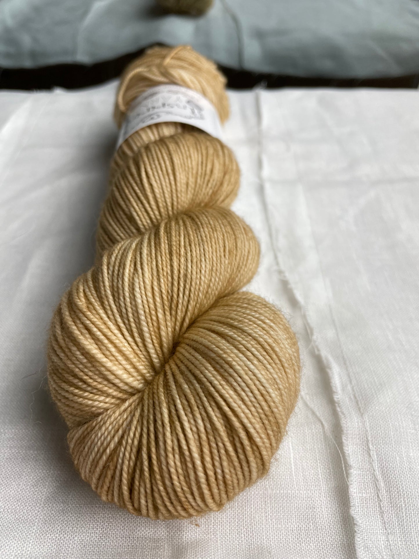 A close up of a skein of tonal golden cream coloured hand dyed Merino Sport yarn branded Inspiring Yarns on a background of white linen.
