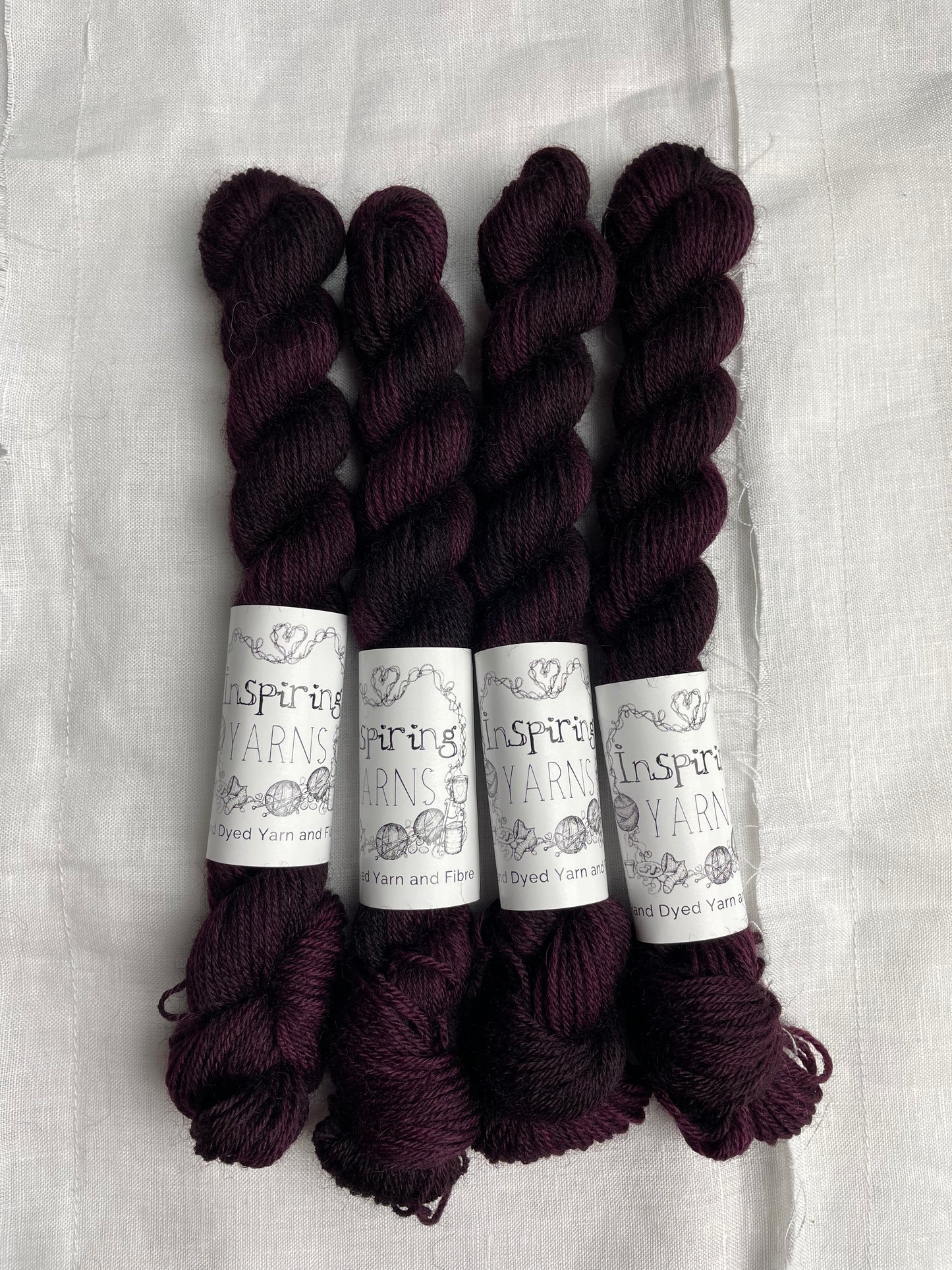Four skeins of 20 gram dark burgundy coloured Highland 4Ply labelled Inspiring Yarns on a white linen background.