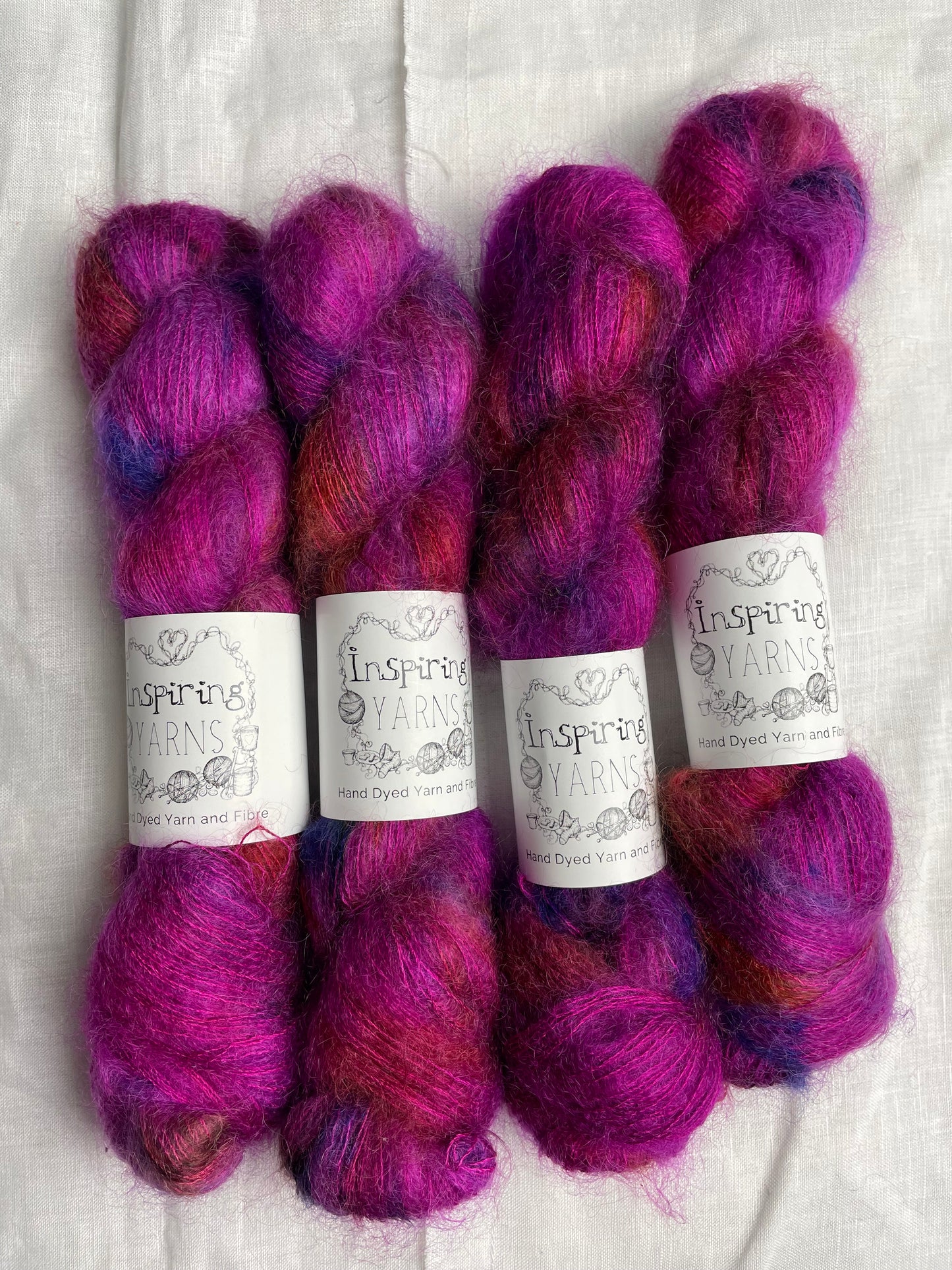 Four skeins of fuchsia pink speckled kid mohair silk lace yarn labelled Inspiring Yarns on a white linen background.