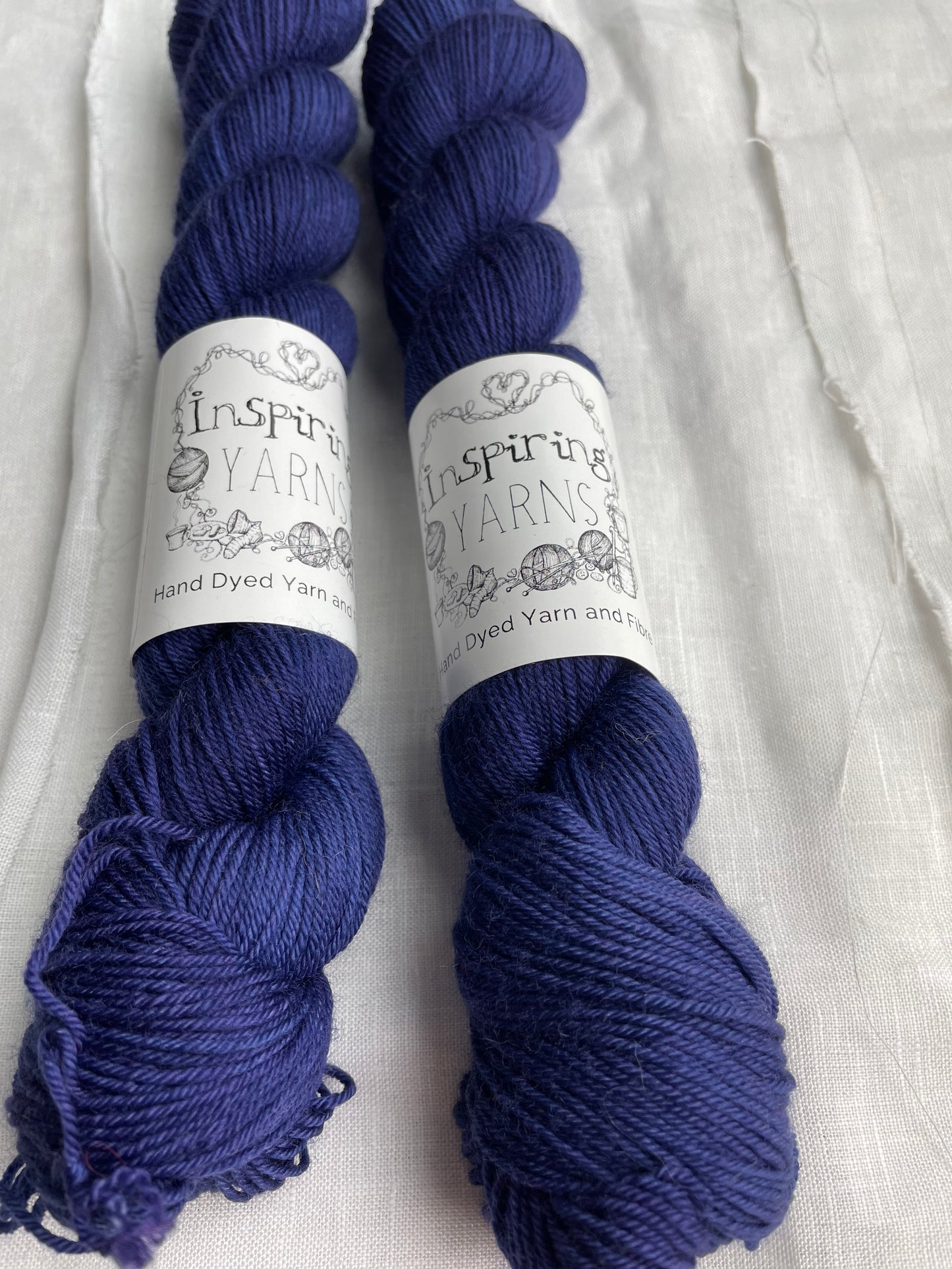 Close up of two tonal deep blue purple coloured skeins of hand dyed Merino Smooth Sock yarn branded Inspiring Yarns on white linen fabric.