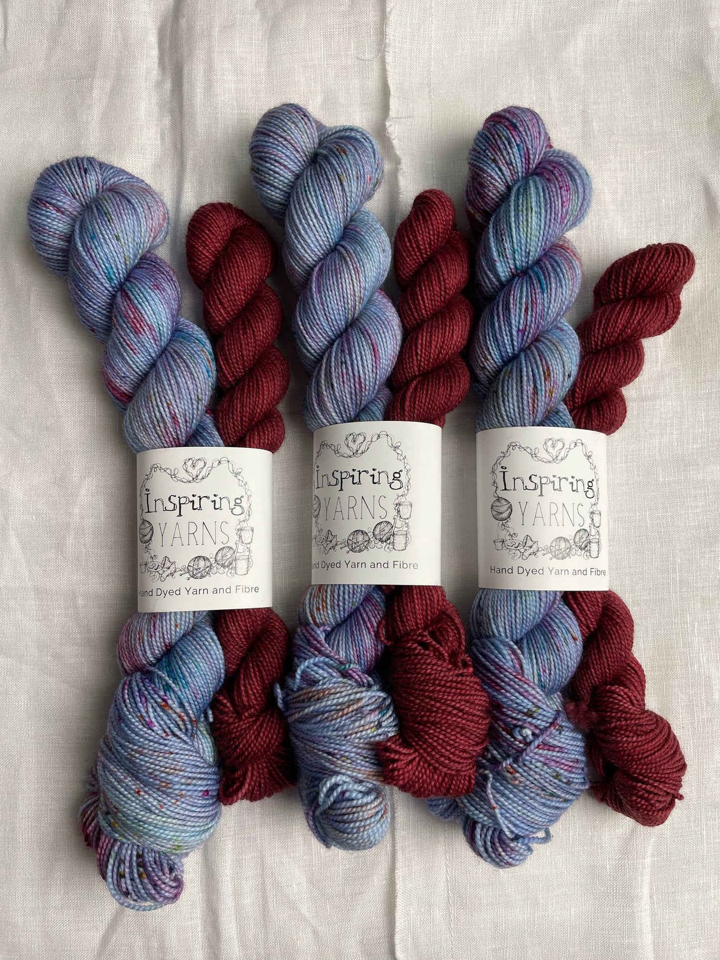 three sock sets made of a skein of light blue speckled hand dyed yarn with a contrasting deep blood red mini skein, labelled Inspiring Yarns and laid on a white linen cloth.