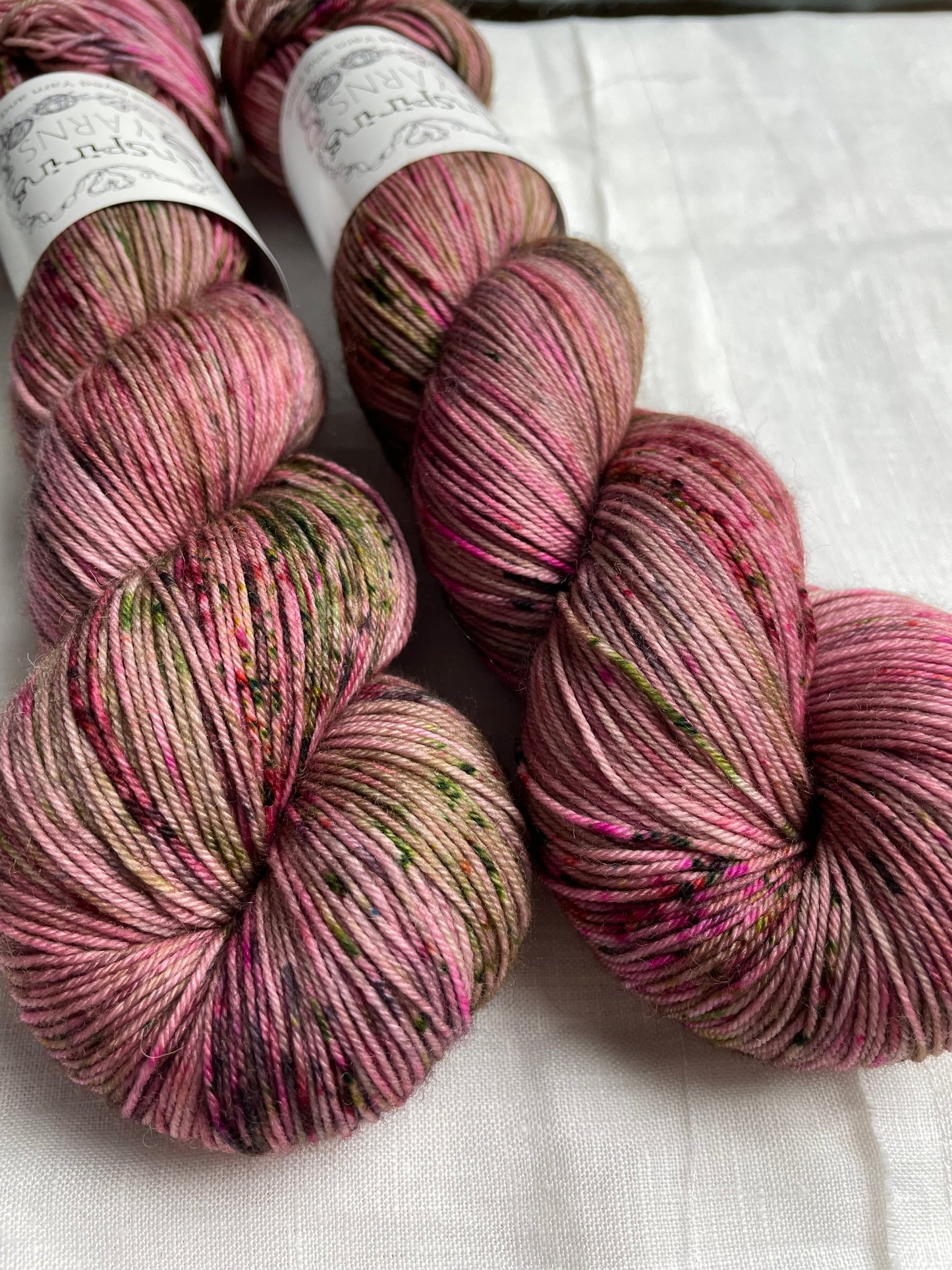 Close up of two speckled pink coloured skeins of Merino Smooth Sock yarn branded Inspiring Yarns on a white linen fabric background.